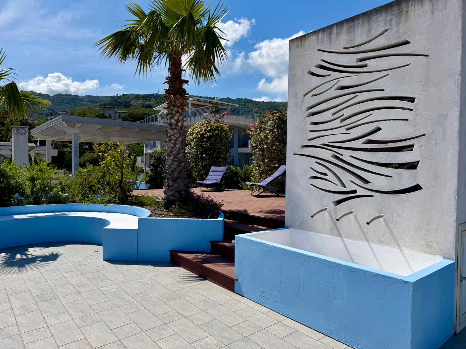 Pannello decorativo in pietra con fontana moderna e piscina all'aperto in un giardino tropicale soleggiato, ideale per relax e svago.