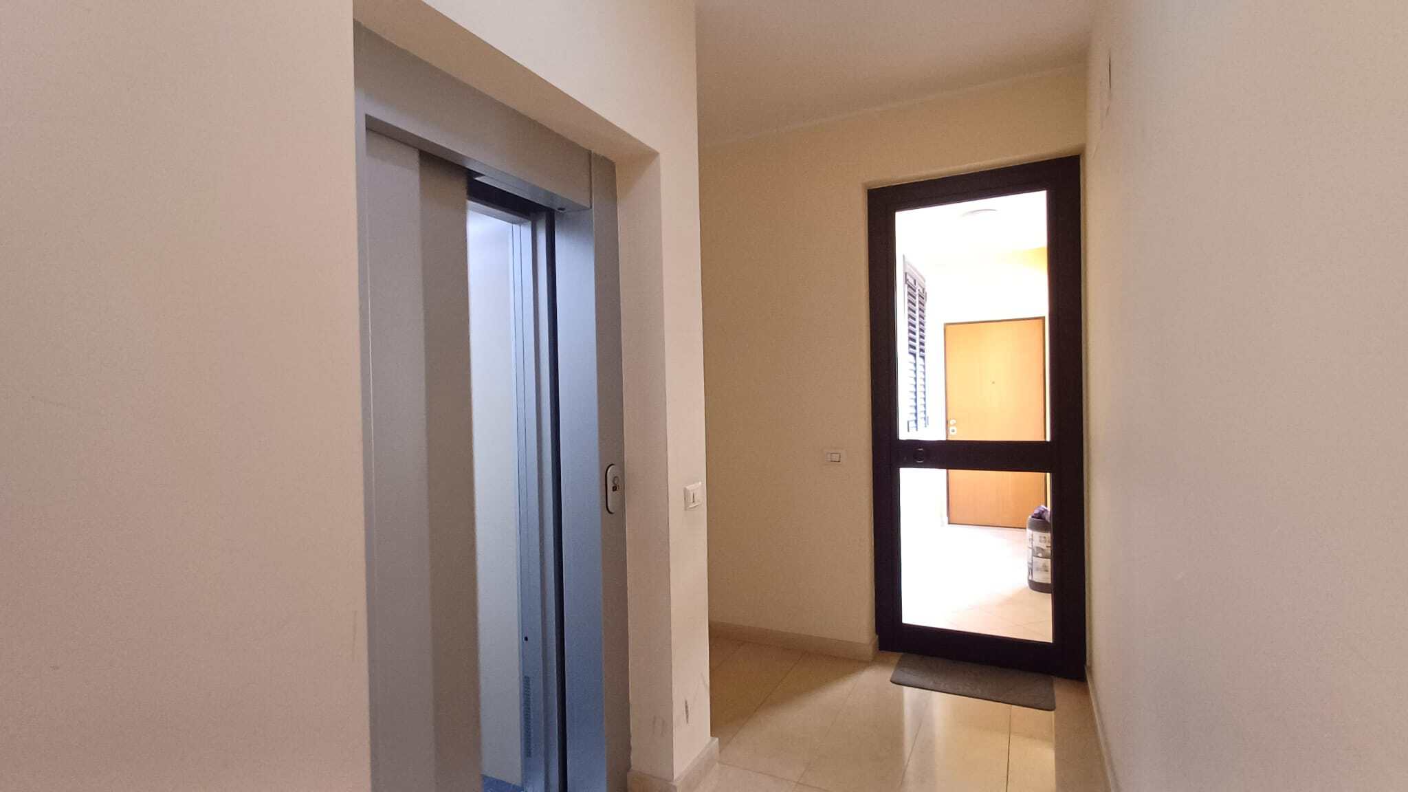 Ascensore e porta finestra che dà accesso a un balcone in un appartamento moderno. Interno luminoso con pareti chiare e pavimento in piastrelle beige, ideale per residenze di qualità.