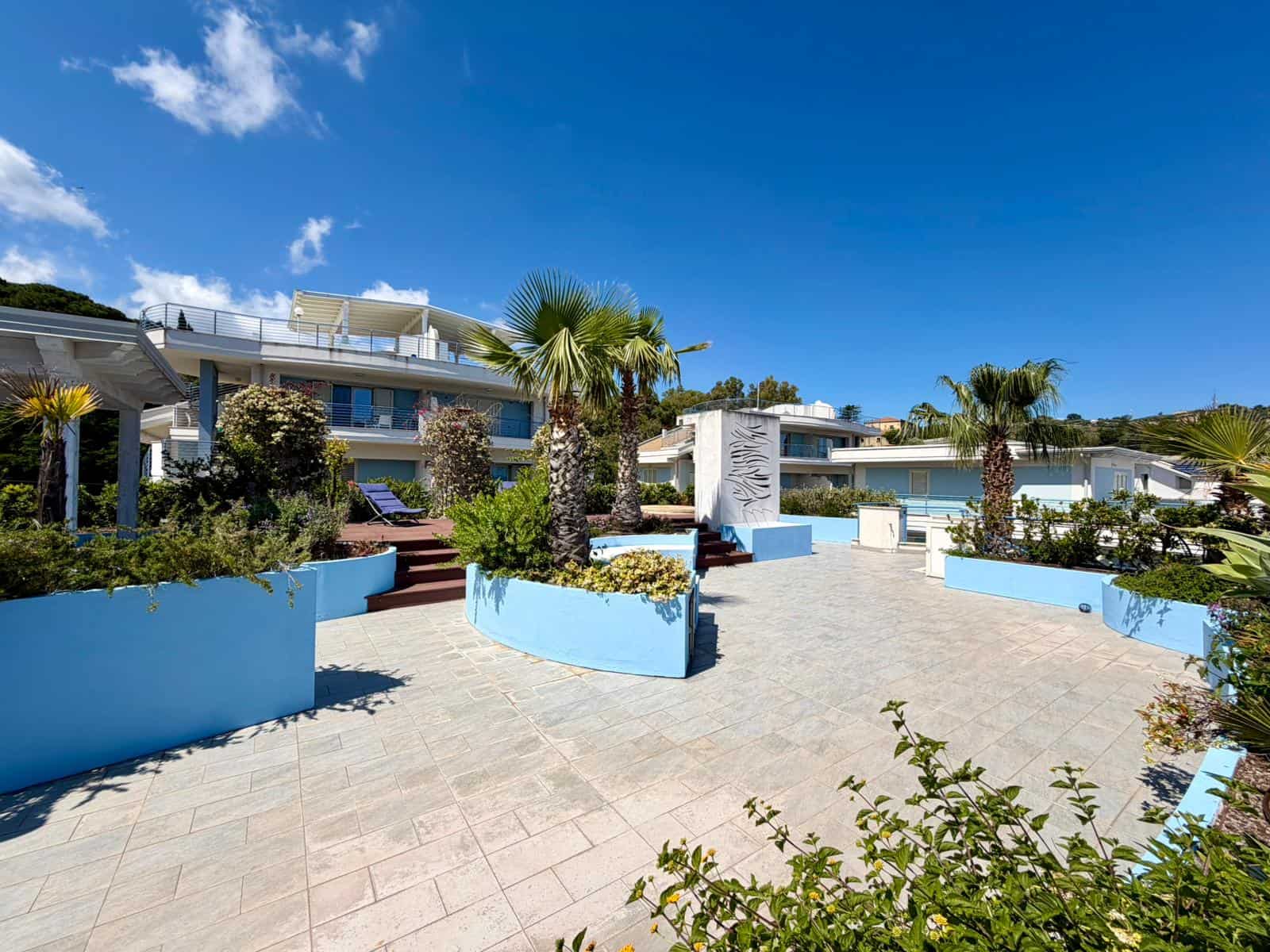 Spazio esterno di una residenza moderna con piante tropicali, pavimentazione in pietra e area relax sotto un cielo sereno, ideale per vivere il comfort e il design contemporaneo.
