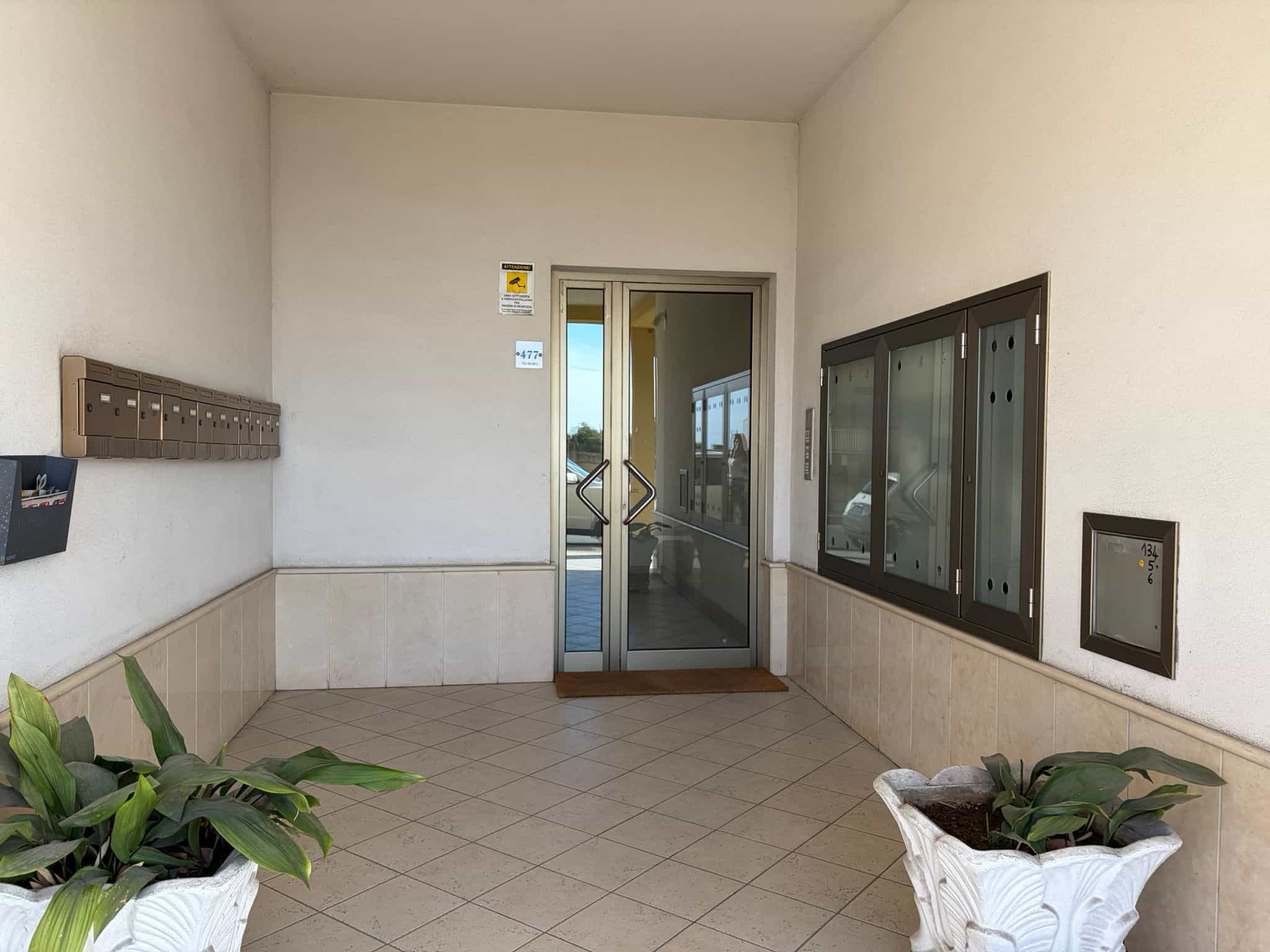 Ingresso condominiale con porta in vetro e piante decorative, ambiente luminoso e accogliente, ideale per l'accesso agli appartamenti.
