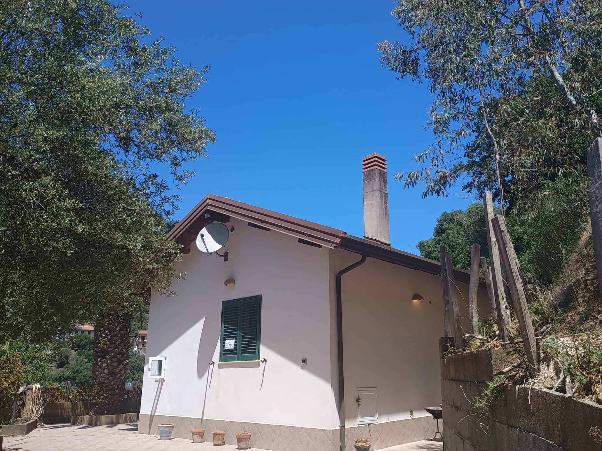 Un'abitazione moderna con pareti bianche, finestra con persiana verde e camino in mattoni, immersa nel verde con un cielo azzurro, ideale per chi cerca una casa accogliente e funzionale.