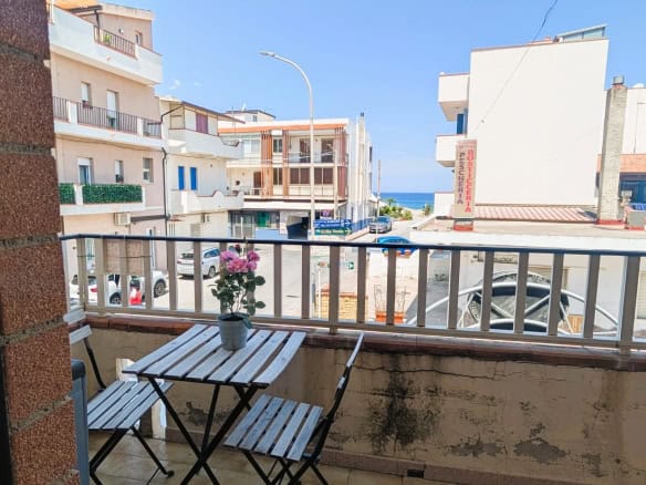Balcone con vista mare, tavolo e sedie in metallo, fiori in vaso, edificio e strada circostante, cielo sereno.