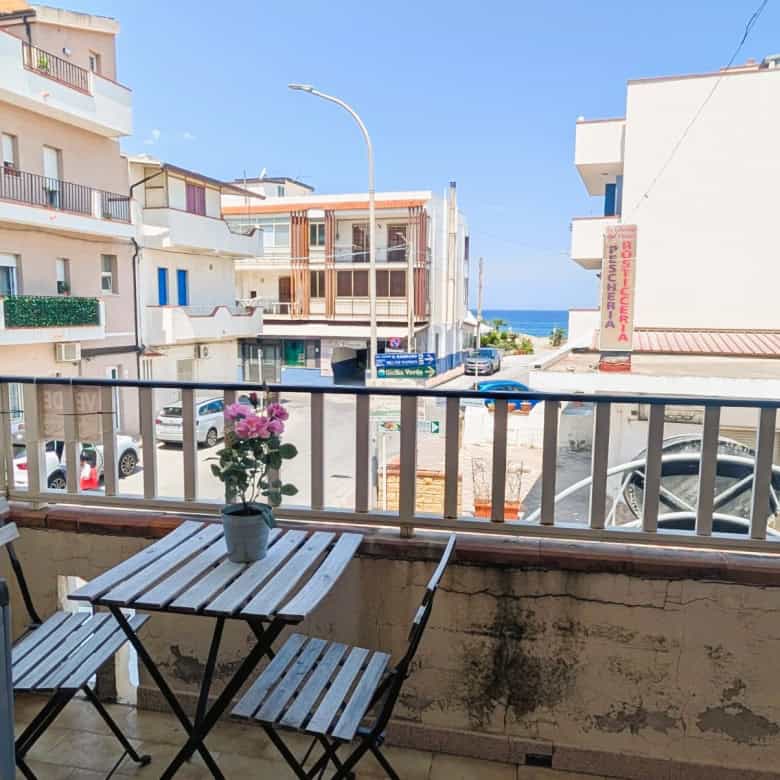 Balcone con vista mare, tavolo e sedie in metallo, fiori in vaso, edificio e strada circostante, cielo sereno.