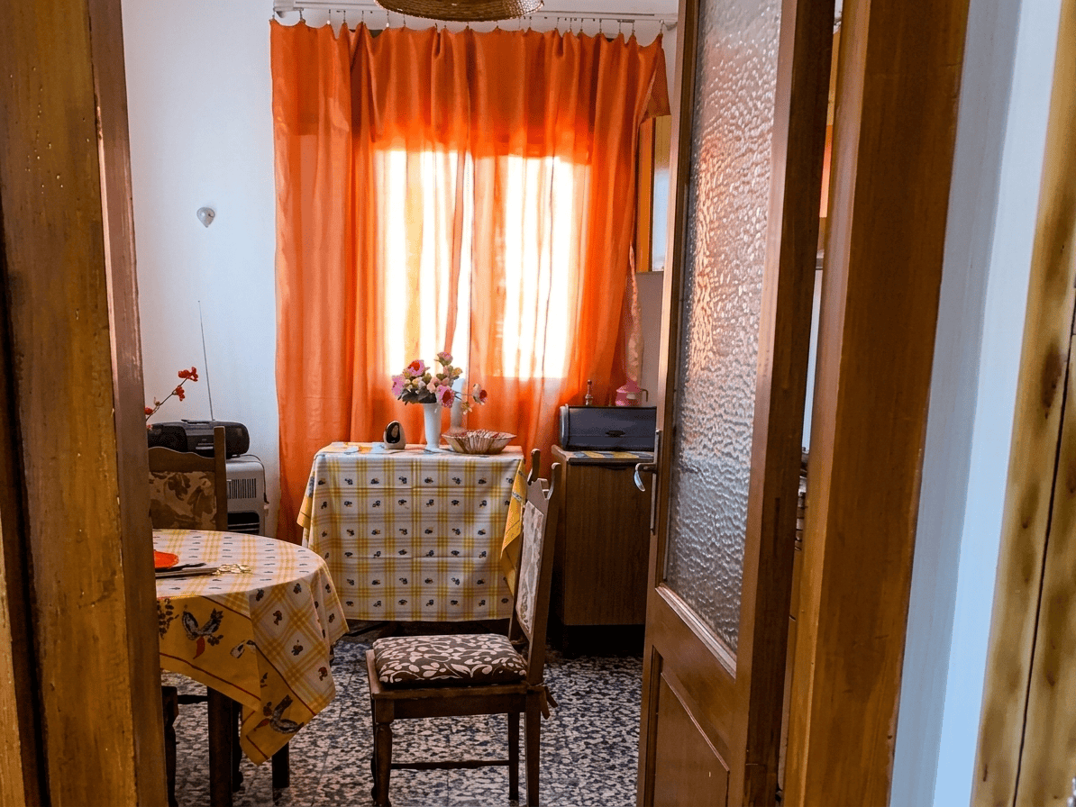 Ambiente interno di una stanza con tende arancioni, tavolo con tovaglia a quadri, sedie e decorazioni floreali, in stile accogliente e luminoso.