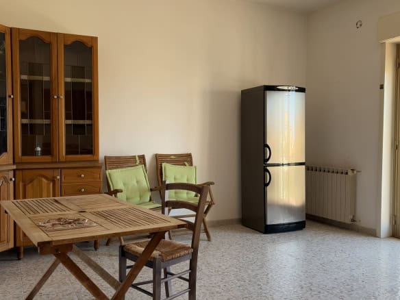 Interno di una sala da pranzo con tavolo, sedie e frigorifero, luce naturale dalla porta finestra.