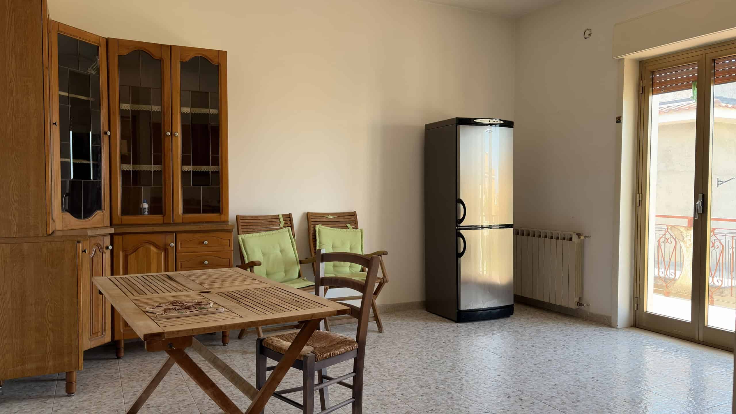 Interno di una sala da pranzo con tavolo, sedie e frigorifero, luce naturale dalla porta finestra.