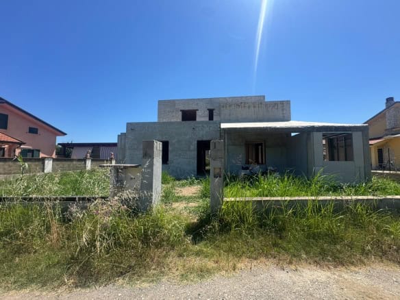 Casa in costruzione con struttura in cemento e giardino in erba alta sotto un cielo azzurro e soleggiato.
