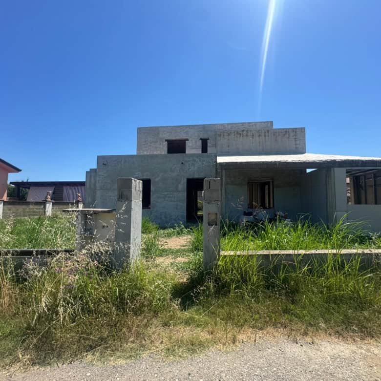 Casa in costruzione con struttura in cemento e giardino in erba alta sotto un cielo azzurro e soleggiato.