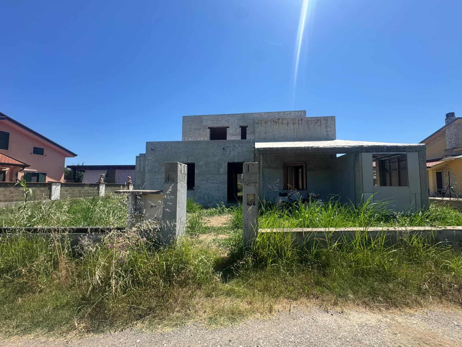 Casa in costruzione con struttura in cemento e giardino in erba alta sotto un cielo azzurro e soleggiato.