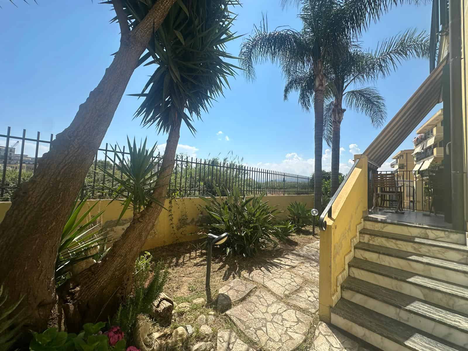 Giardino esterno con piante tropicali e scale che portano a un appartamento in vendita a Milazzo, Sicilia.