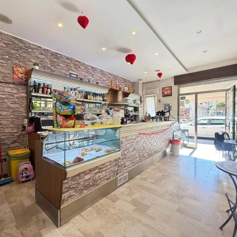 Interno di un bar o caffetteria con bancone in pietra, esposizione di dolci e bevande, decorazioni a cuore rosso, ingresso con vetrata e tavoli.