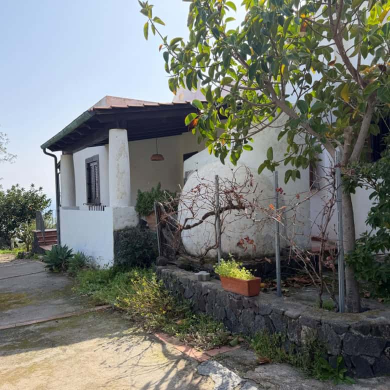Un'abitazione rustica con pareti bianche, tetto in tegole e giardino con piante e alberi, immersa in un ambiente naturale e tranquillo, ideale per una casa di campagna o una seconda casa in campagna.