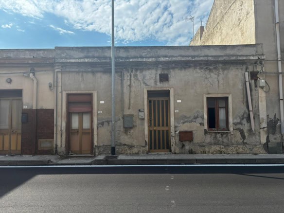 Vecchia casa in stile rustico con pareti in pietra e finestre piccole, situata lungo una strada urbana sotto un cielo sereno, rappresenta un esempio di architettura tradizionale italiana.