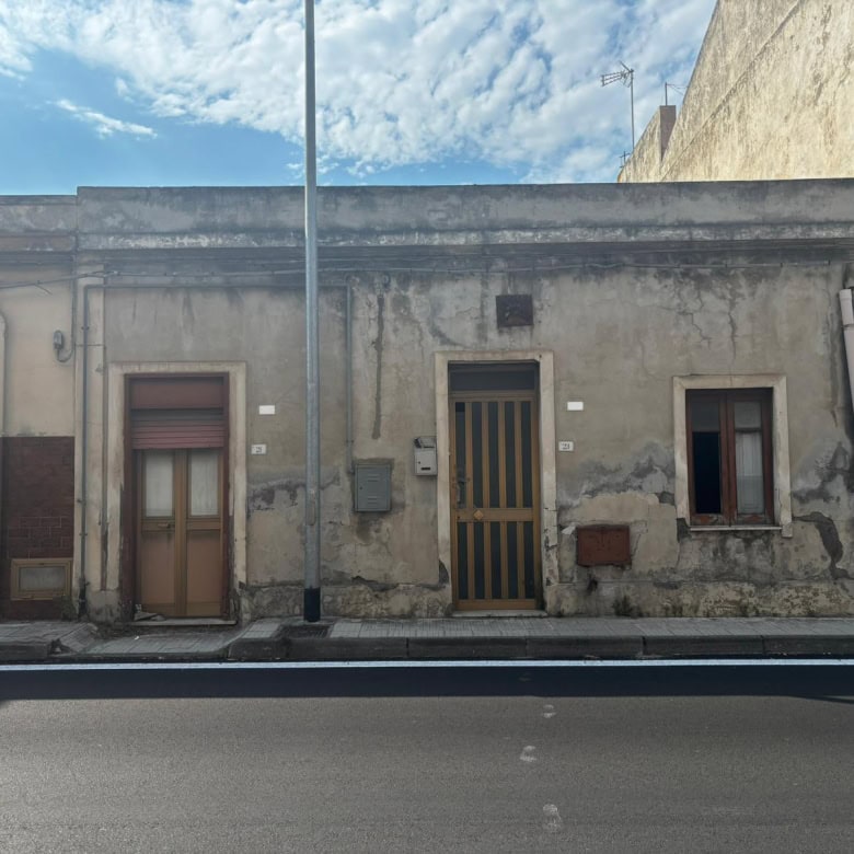 Vecchia casa in stile rustico con pareti in pietra e finestre piccole, situata lungo una strada urbana sotto un cielo sereno, rappresenta un esempio di architettura tradizionale italiana.