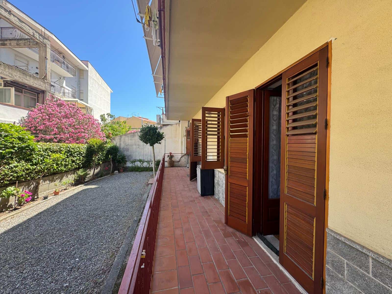 Ampia veranda con persiane in legno, fioriera e giardino esterno soleggiato, ideale per relax e convivialità, in zona residenziale tranquilla.