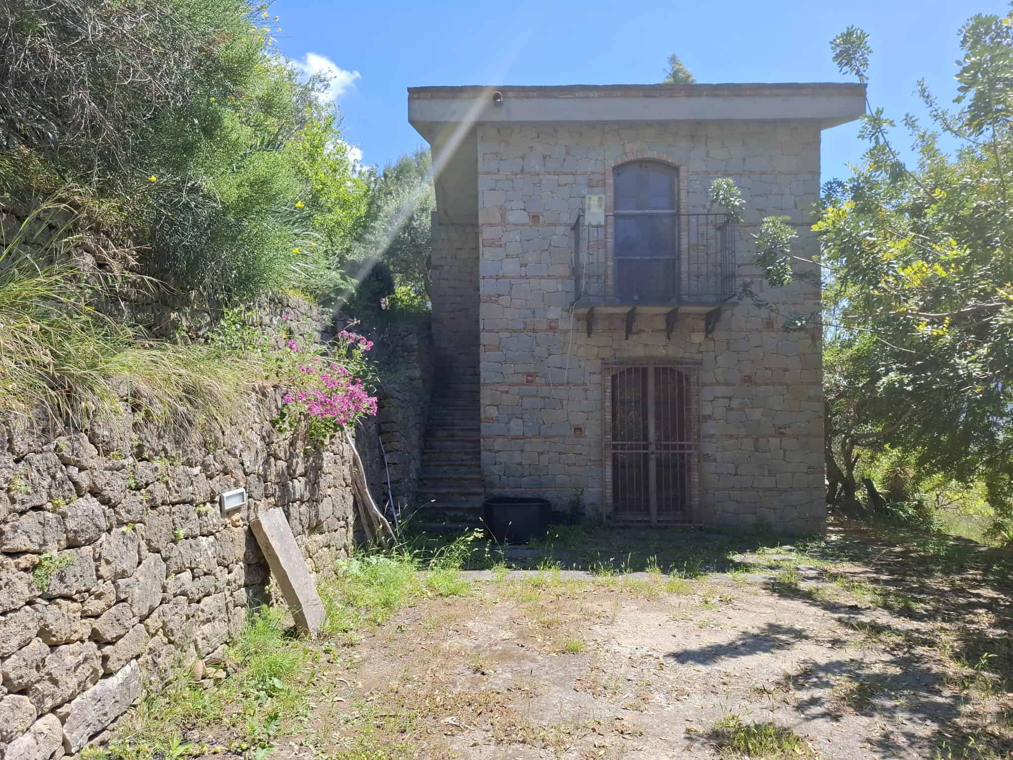 Antico edificio in pietra con balcone e scale esterne, immerso in un ambiente naturale con vegetazione e fiori rosa, ideale per investimento immobiliare o ristrutturazione.