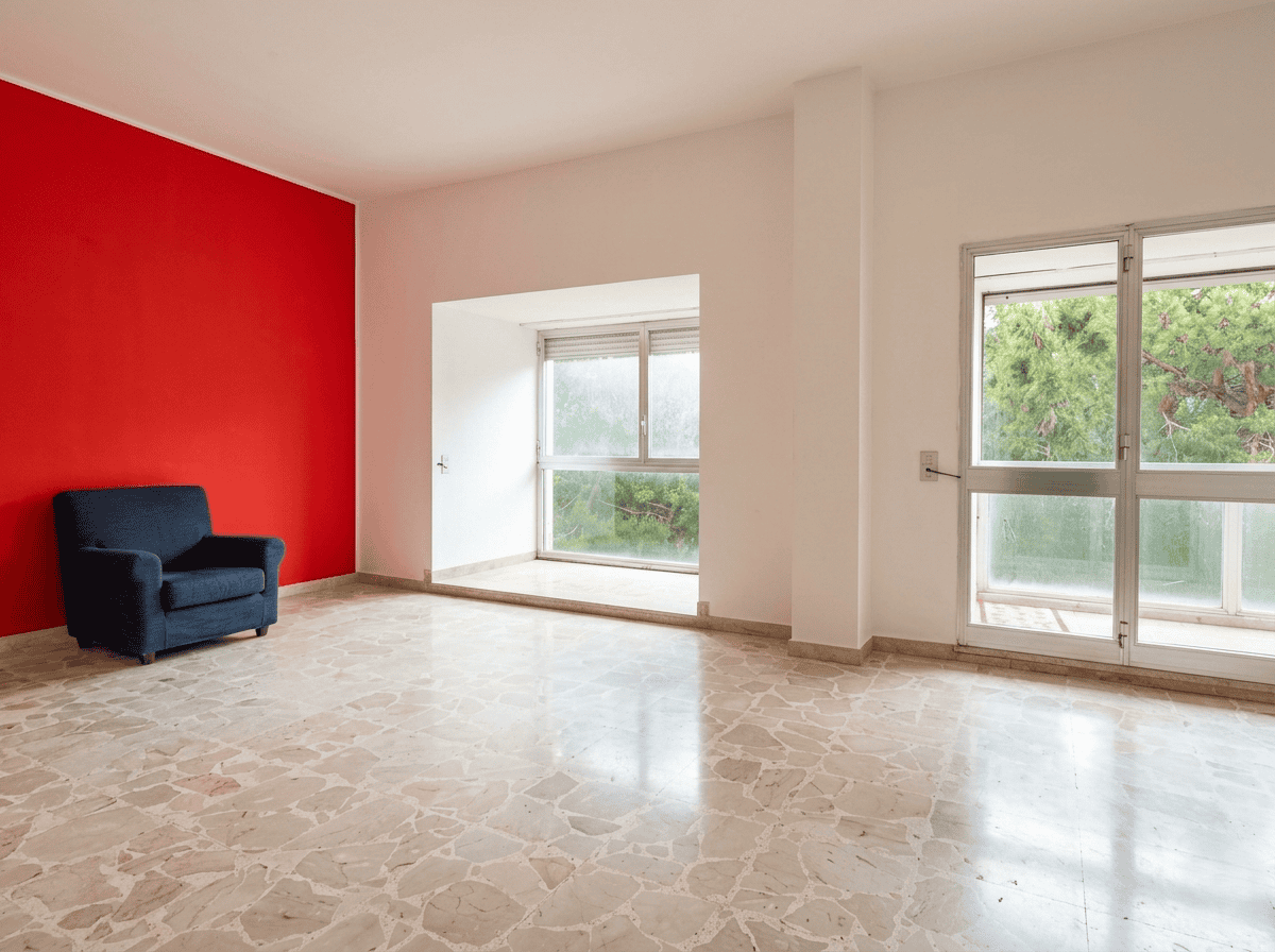 Appartamento luminoso con grandi finestre e parete rossa, ideale per ambienti di soggiorno o studio. Spazio aperto, pavimento in marmo e vista sulla natura circostante.