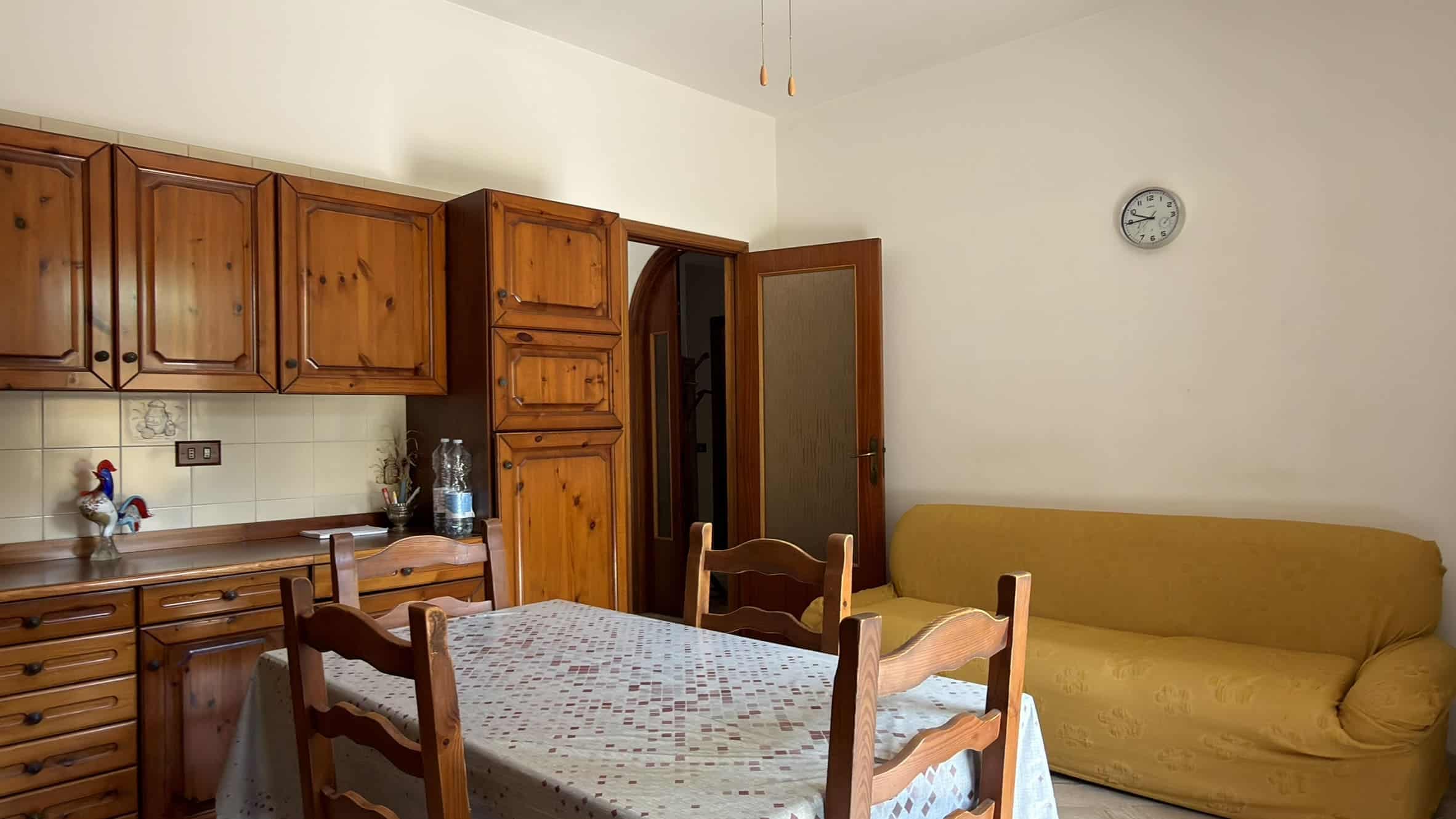 Interno di una cucina con mobili in legno, tavolo con sedie e divano giallo, ambiente luminoso e accogliente.