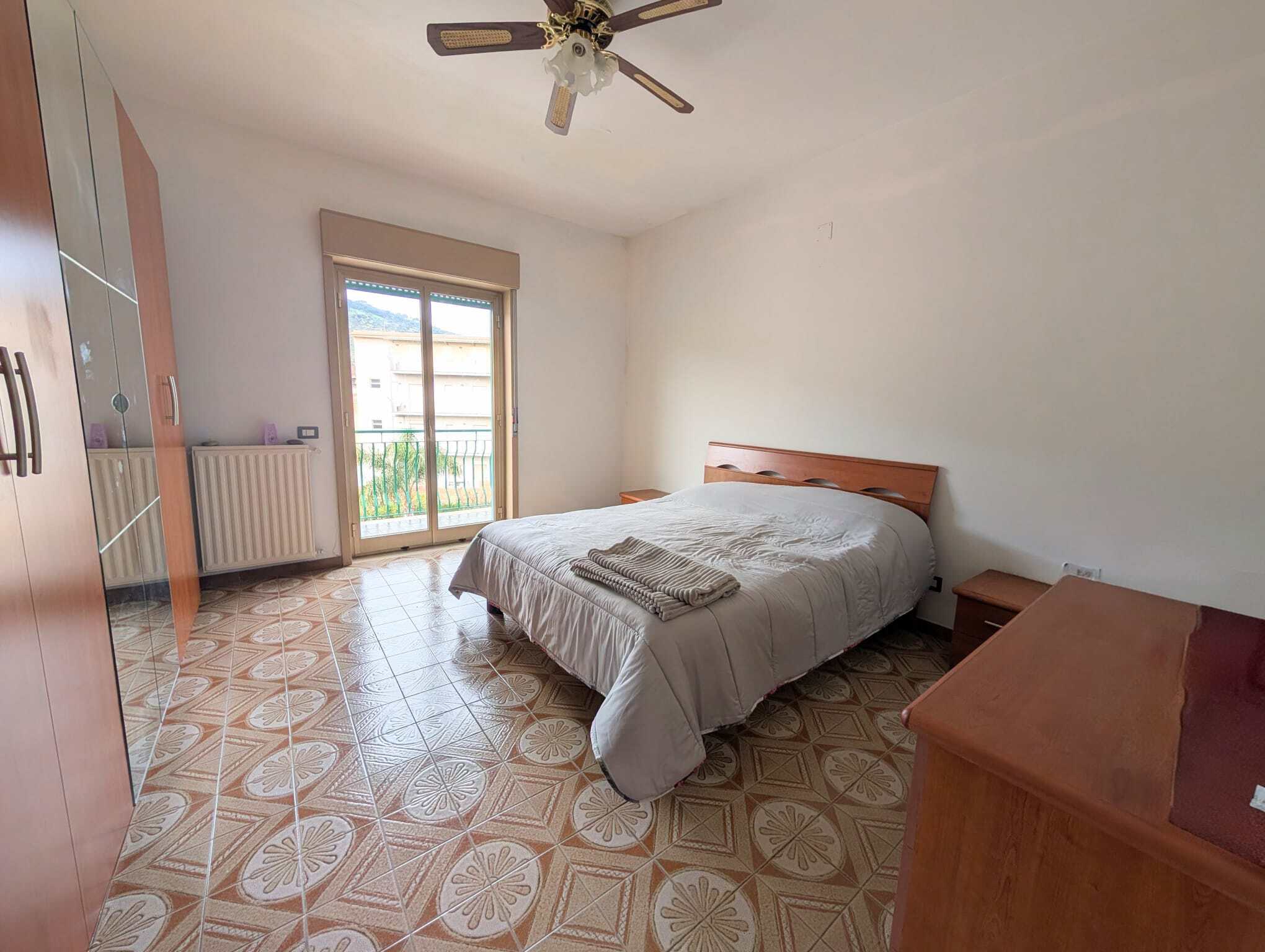 Camera rivolta verso una camera da letto luminosa con letto matrimoniale, armadio, comodini e balcone con vista.