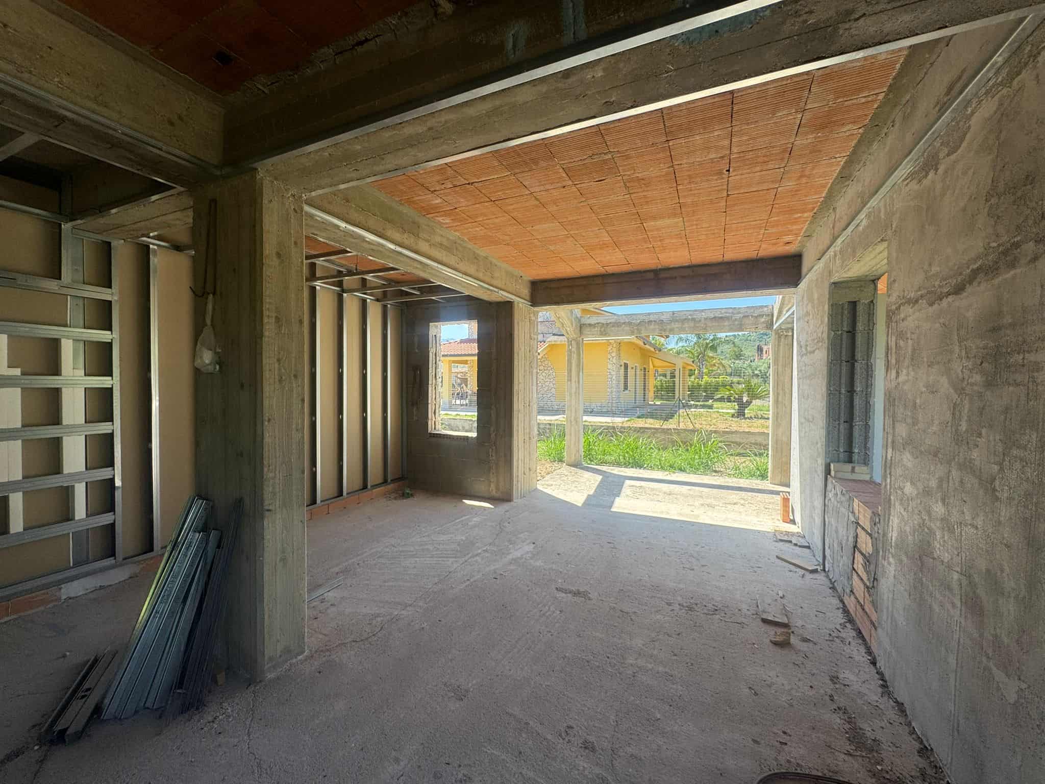 Interno di una casa in costruzione con pareti in cemento e grandi finestre, vista sul giardino esterno.