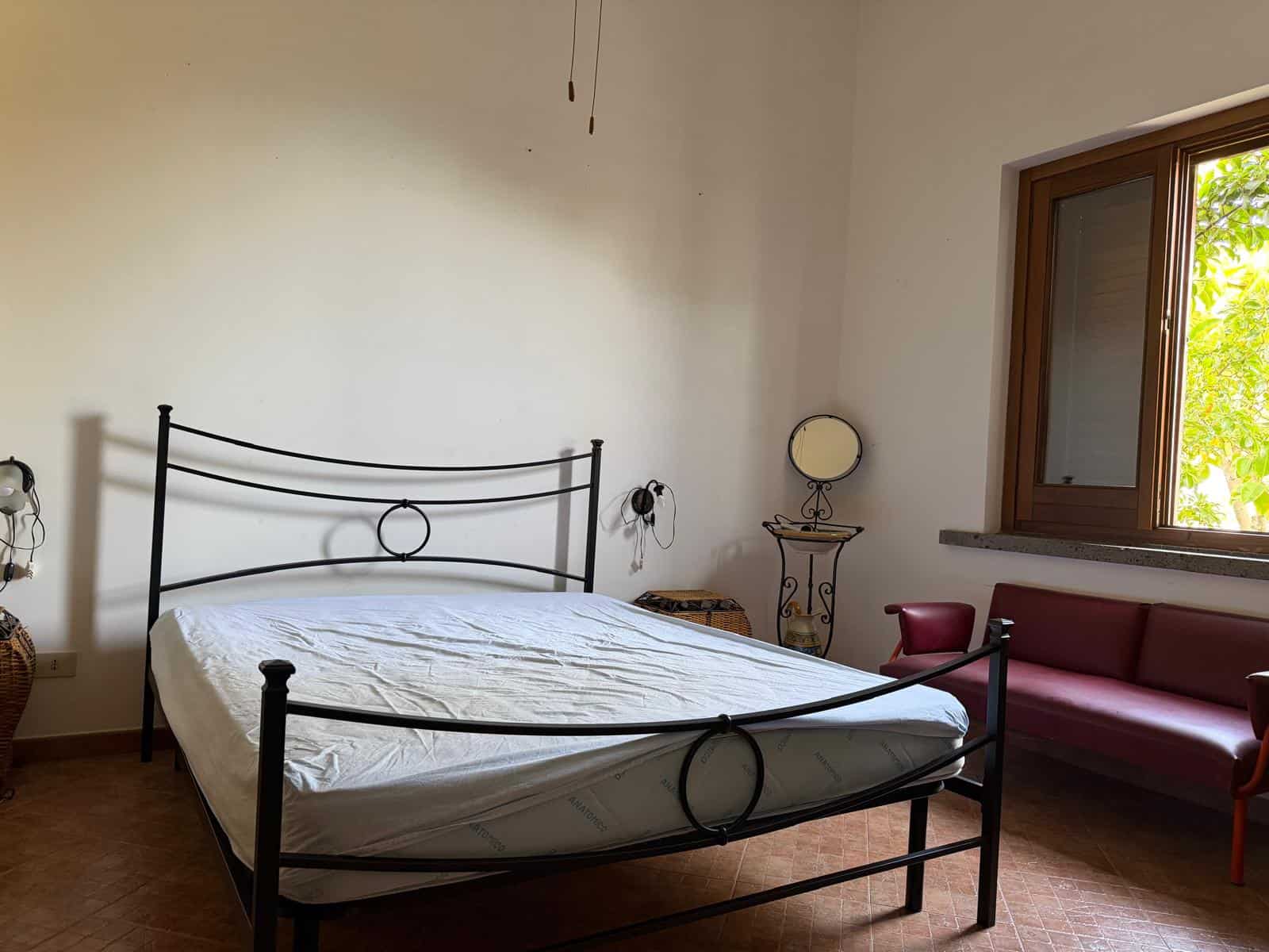 Letto matrimoniale con testiera in metallo nero, in una stanza luminosa con pareti bianche, finestra con vista su alberi verdi, e arredi semplici e funzionali.