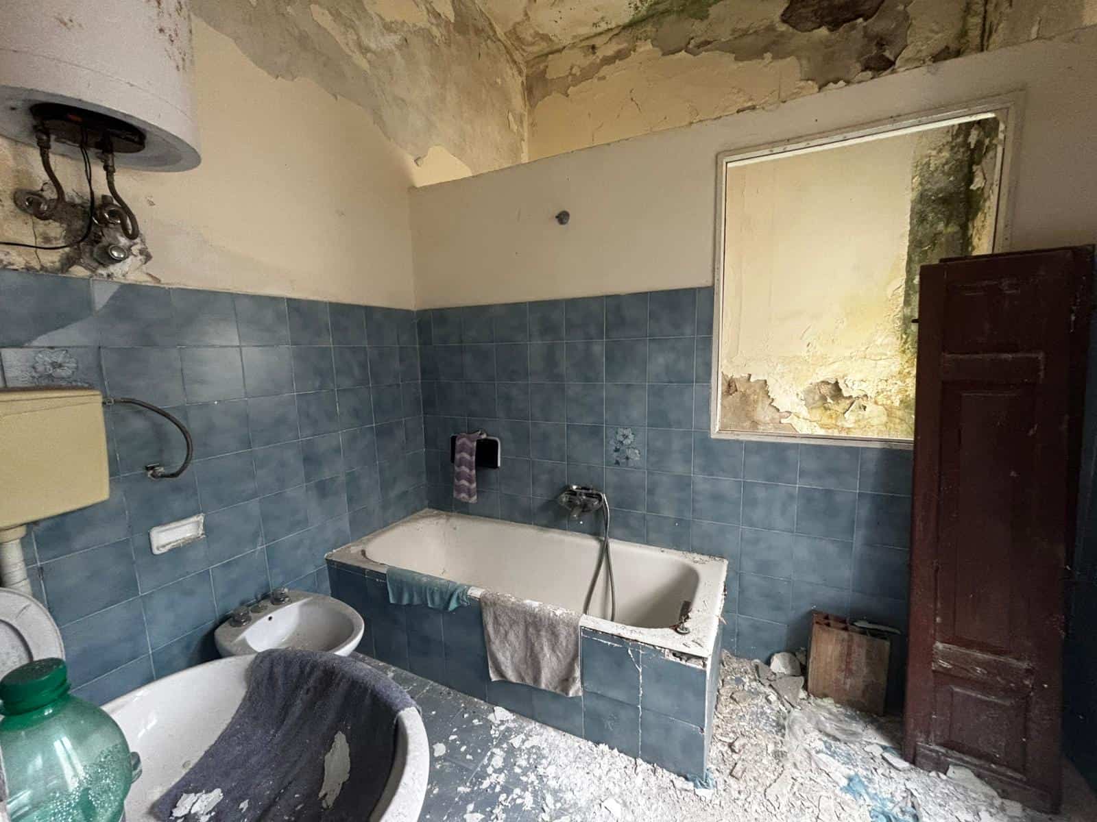 Interno di bagno abbandonato con vasca, pareti in piastrelle blu e soffitto deteriorato, ambiente trascurato e in stato di degrado.