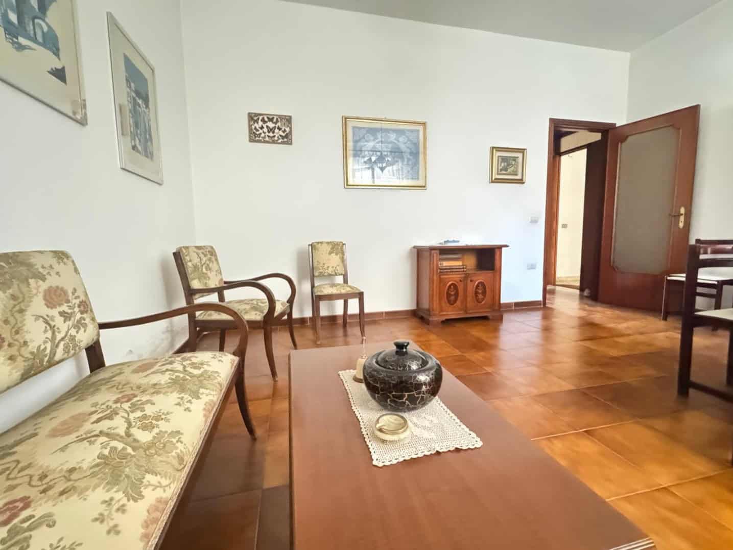 Soggiorno spazioso con mobili in legno, divani e sedie con rivestimenti floreali, pareti decorate con quadri e un pavimento in parquet, ideale per vivere momenti di relax e convivialità.