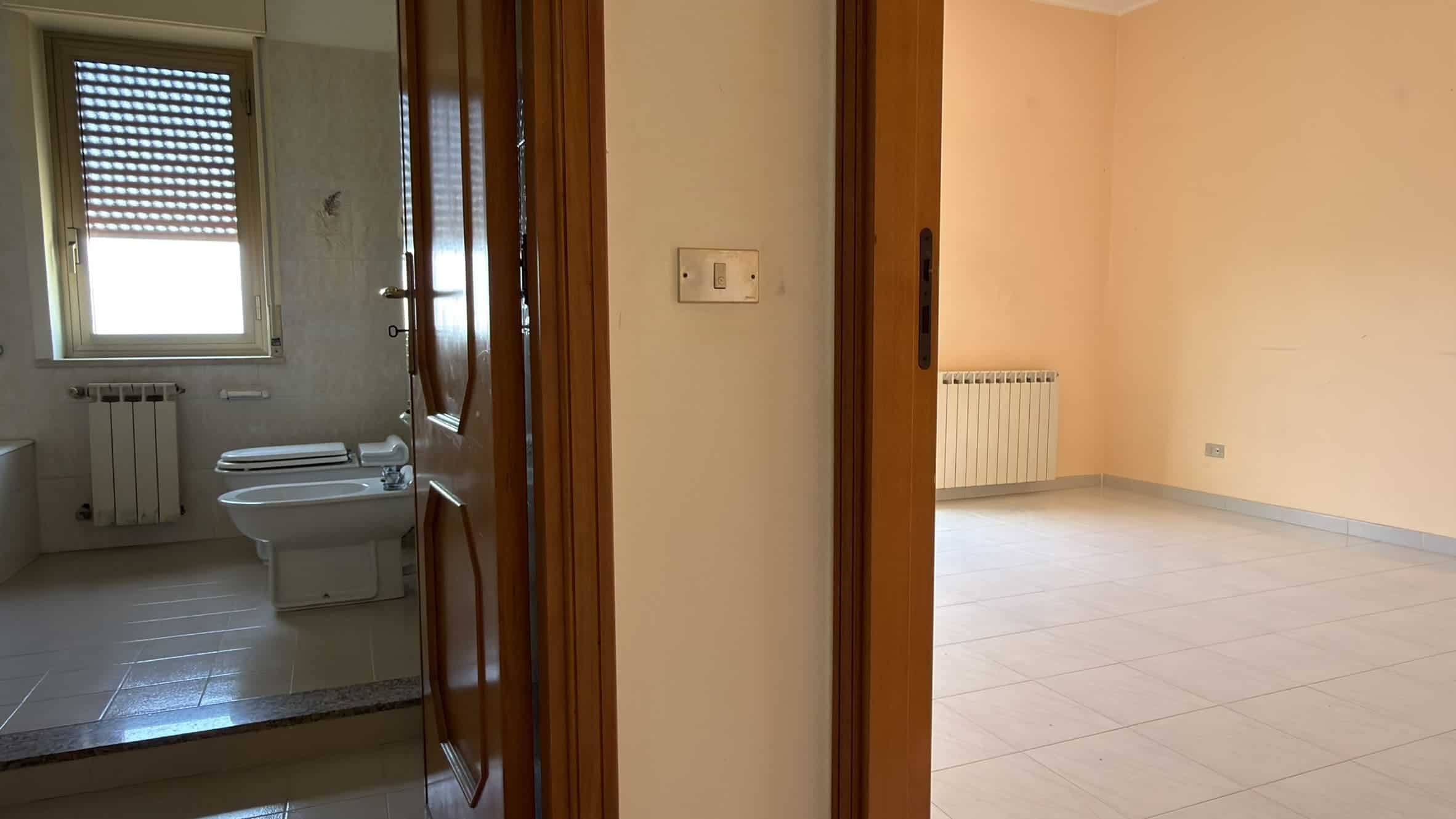 Interno di un appartamento con bagno visibile attraverso una porta aperta, pareti neutre e pavimento in piastrelle chiare.