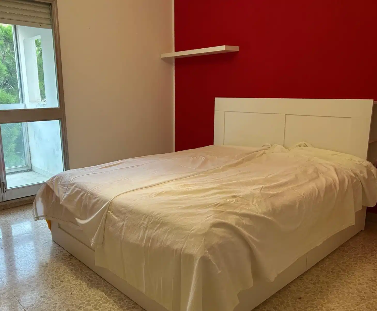Letto matrimoniale in una camera moderna con parete rossa e finestra che lascia entrare luce naturale, arredamento minimalista, ideale per appartamenti in affitto o vendita.