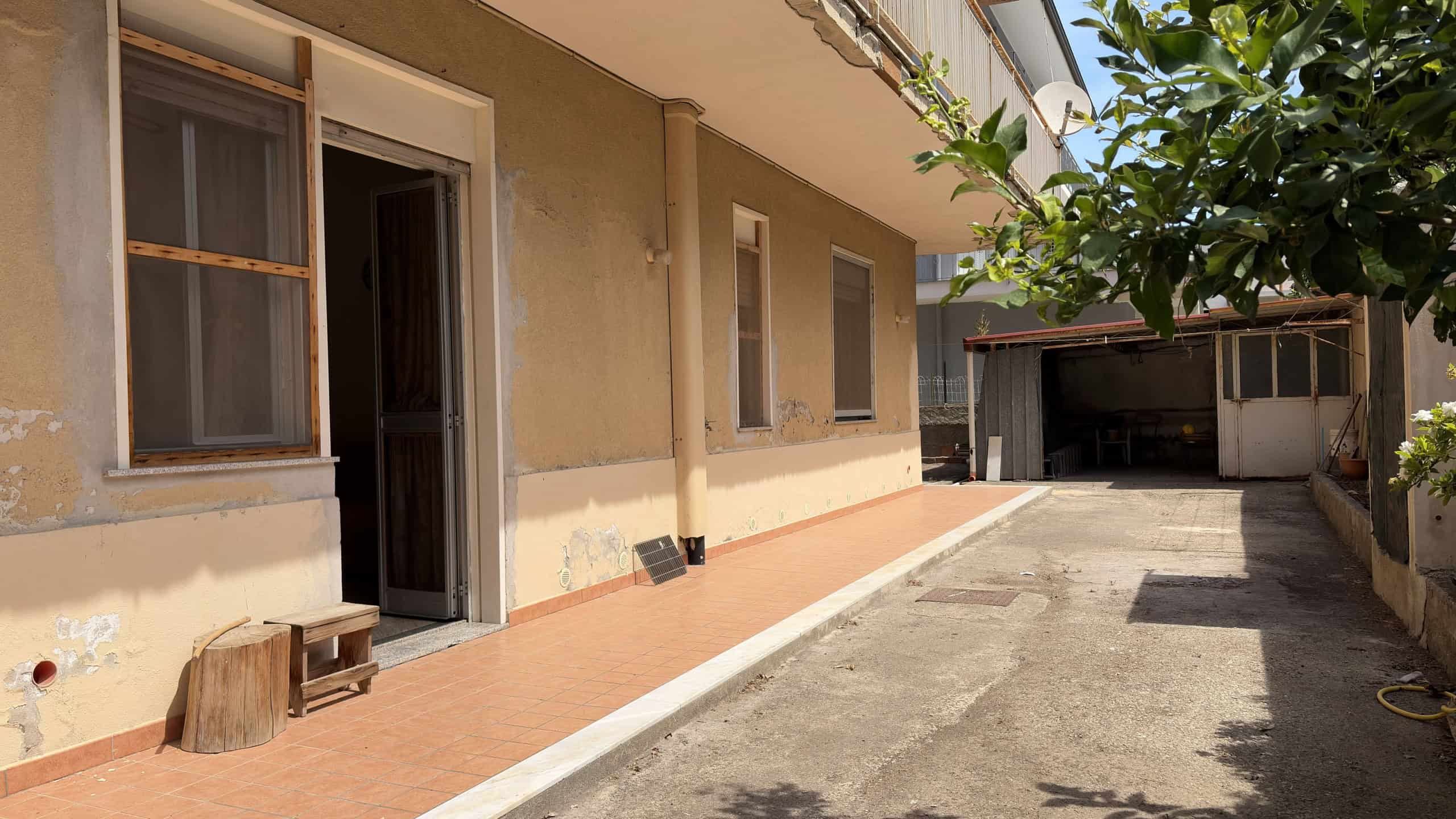 Esterno di una casa con cortile, pareti beige, finestre e porta, area pavimentata e piccolo deposito in fondo.