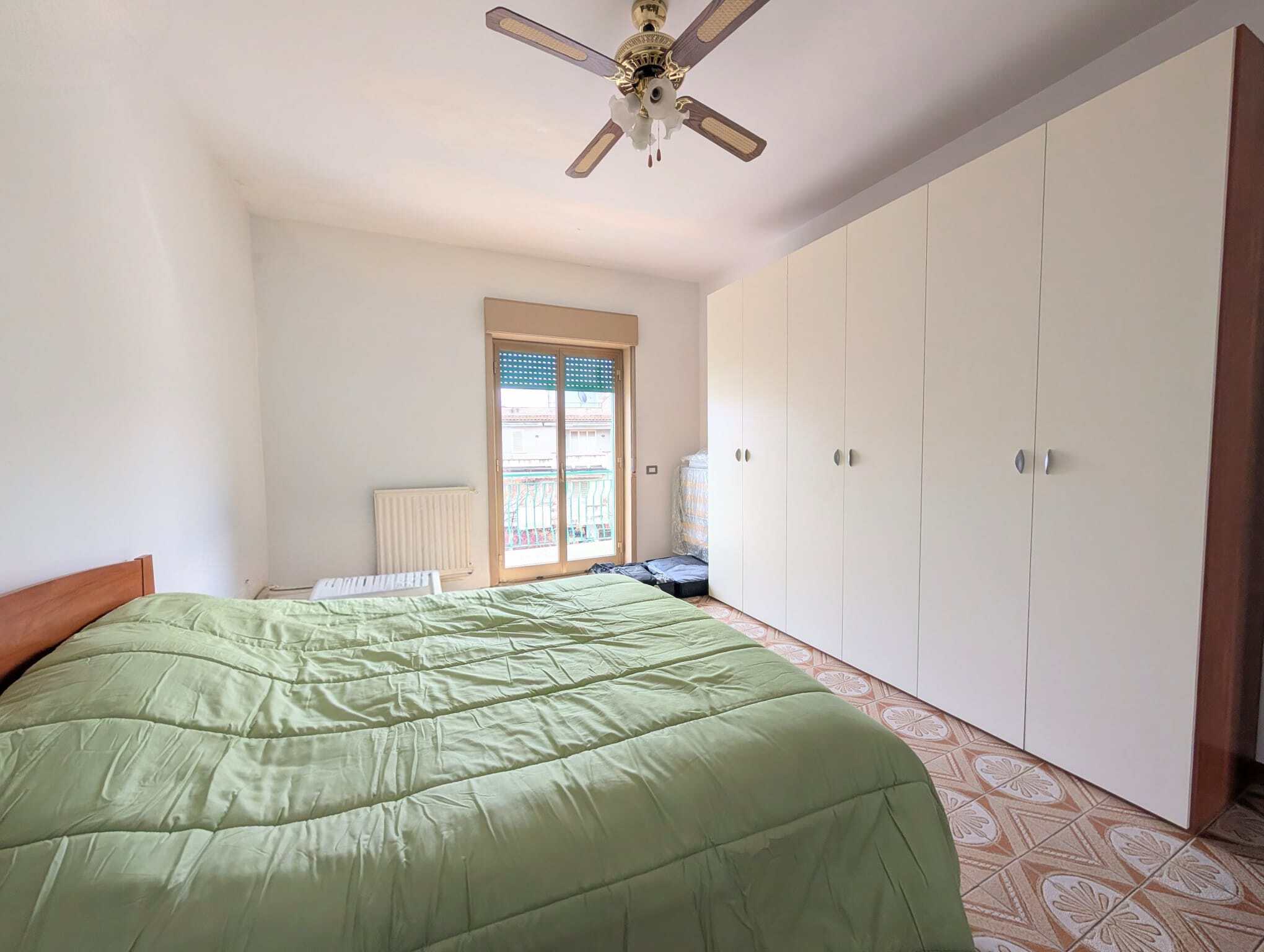 Camera che mostra una camera da letto luminosa con letto matrimoniale, armadio bianco e finestra con vista su un edificio vicino.