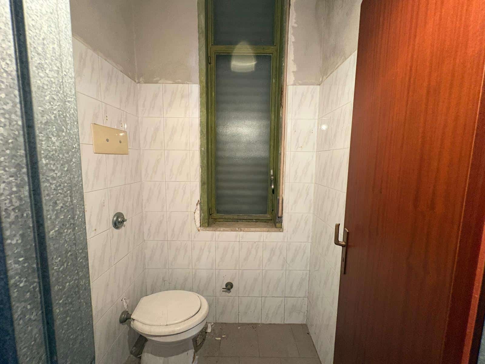 Bagno piccolo con finestra, WC e pareti rivestite in piastrelle bianche.