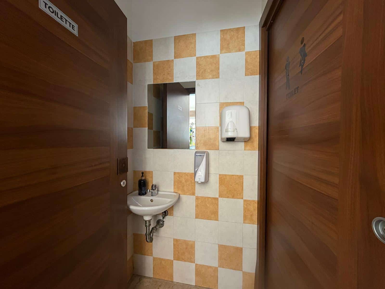 Bagno pubblico con lavabo, specchio e dispenser di sapone, in un ambiente con pareti a scacchi beige e arancioni.