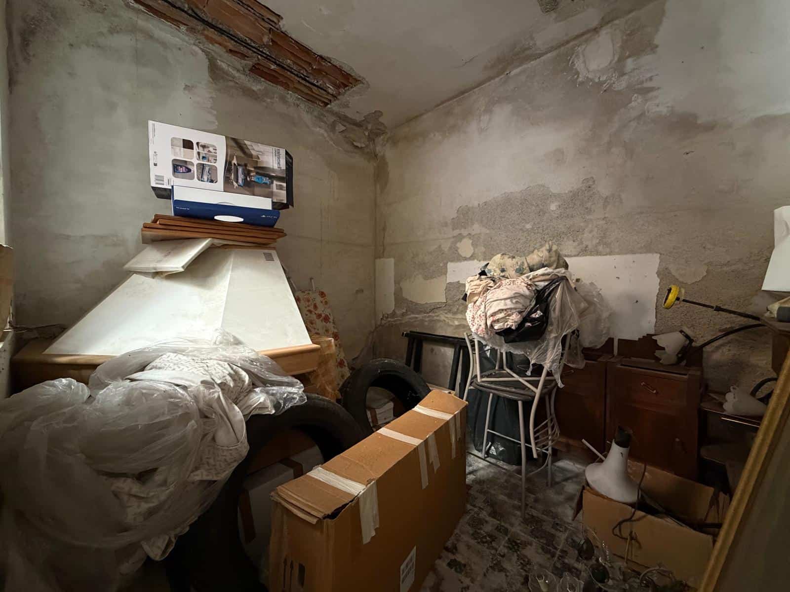 Interno di una casa in fase di ristrutturazione con materiali e mobili impilati, pareti grezze e soffitto con mattoni a vista.