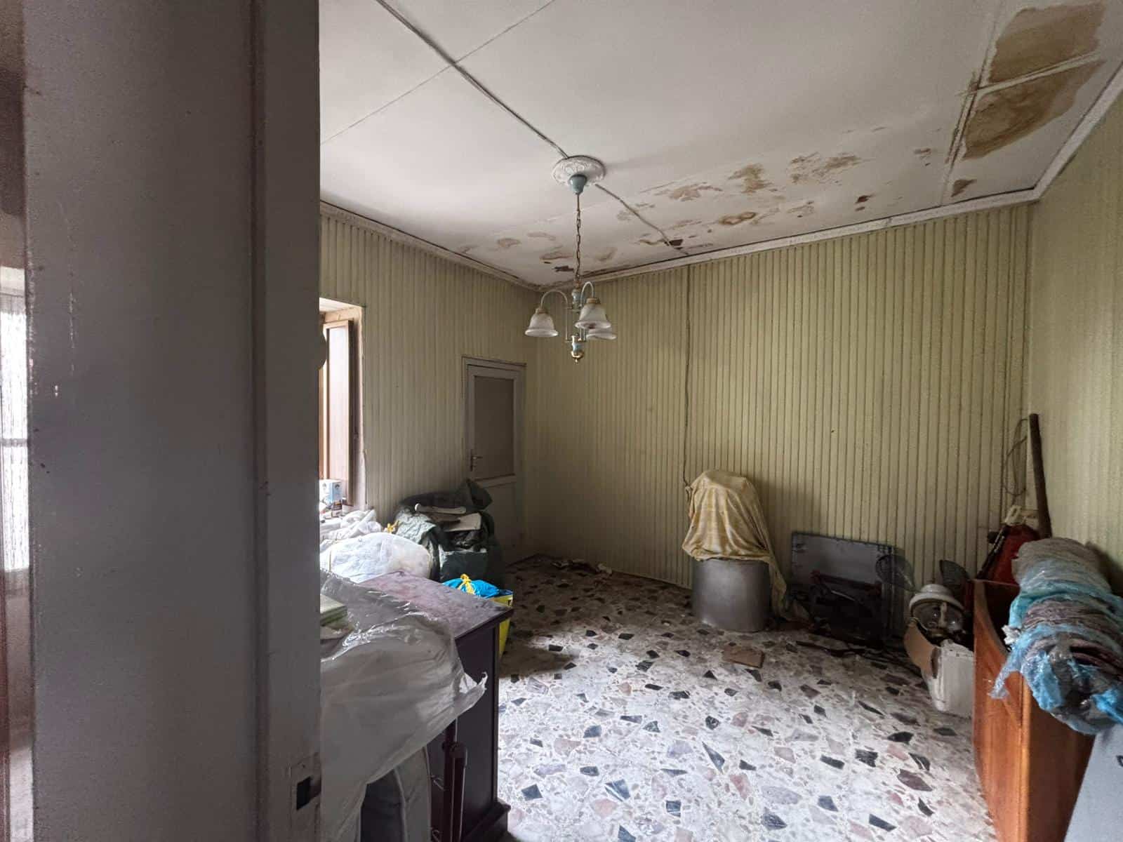 Interno di una stanza abbandonata con soffitto danneggiato e mobili disordinati, ambiente trascurato e in stato di degrado.