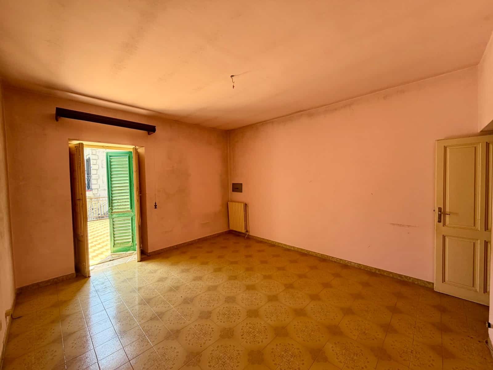 Un ambiente interno vuoto con pareti rosa pallido, pavimento in piastrelle beige e una porta finestra con persiane verdi che permette l'ingresso di luce naturale.