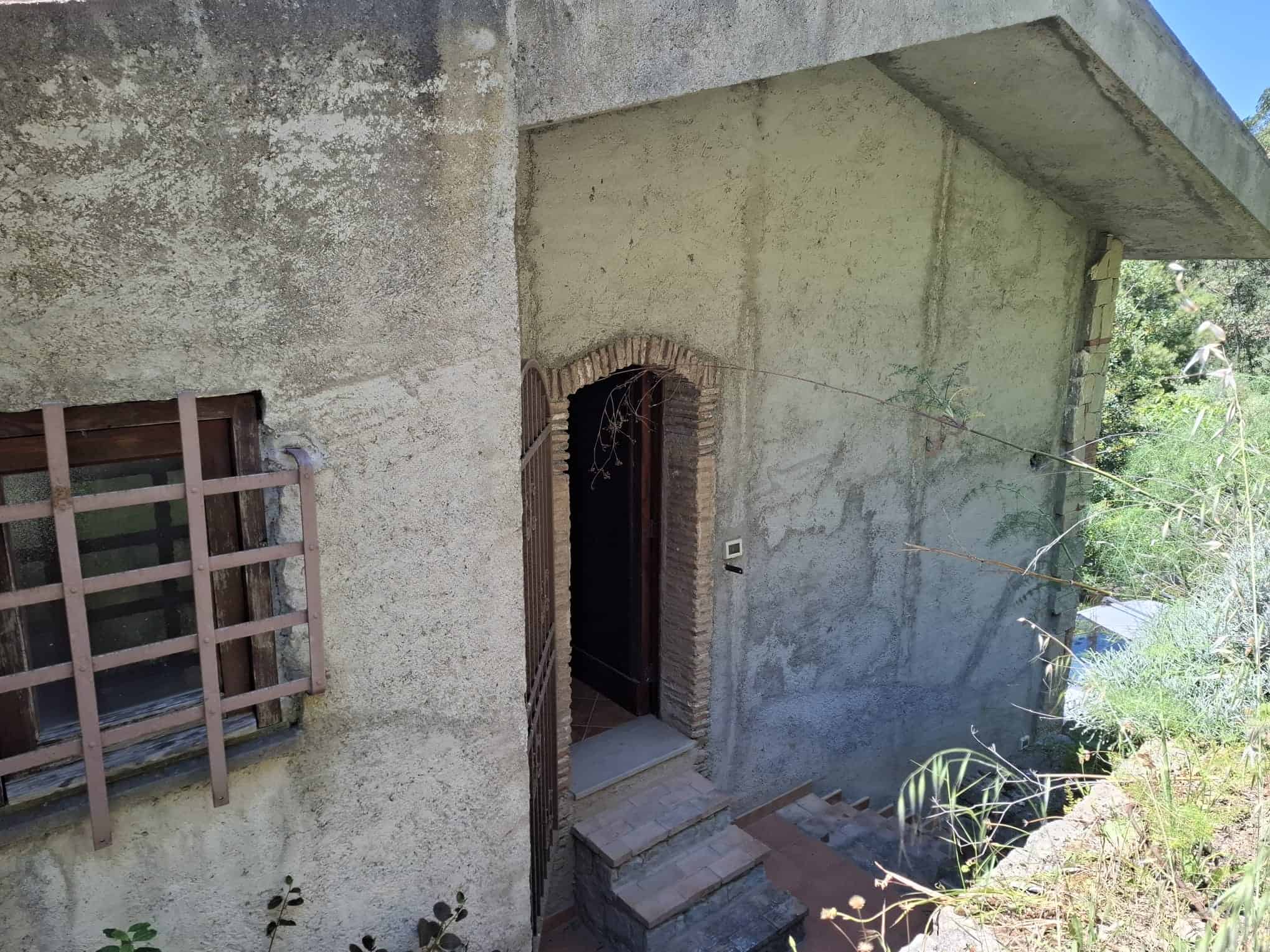 Un ingresso di una casa in stile rustico con pareti in cemento e arco in mattoni, circondato da vegetazione verde e piante, in un ambiente esterno soleggiato.