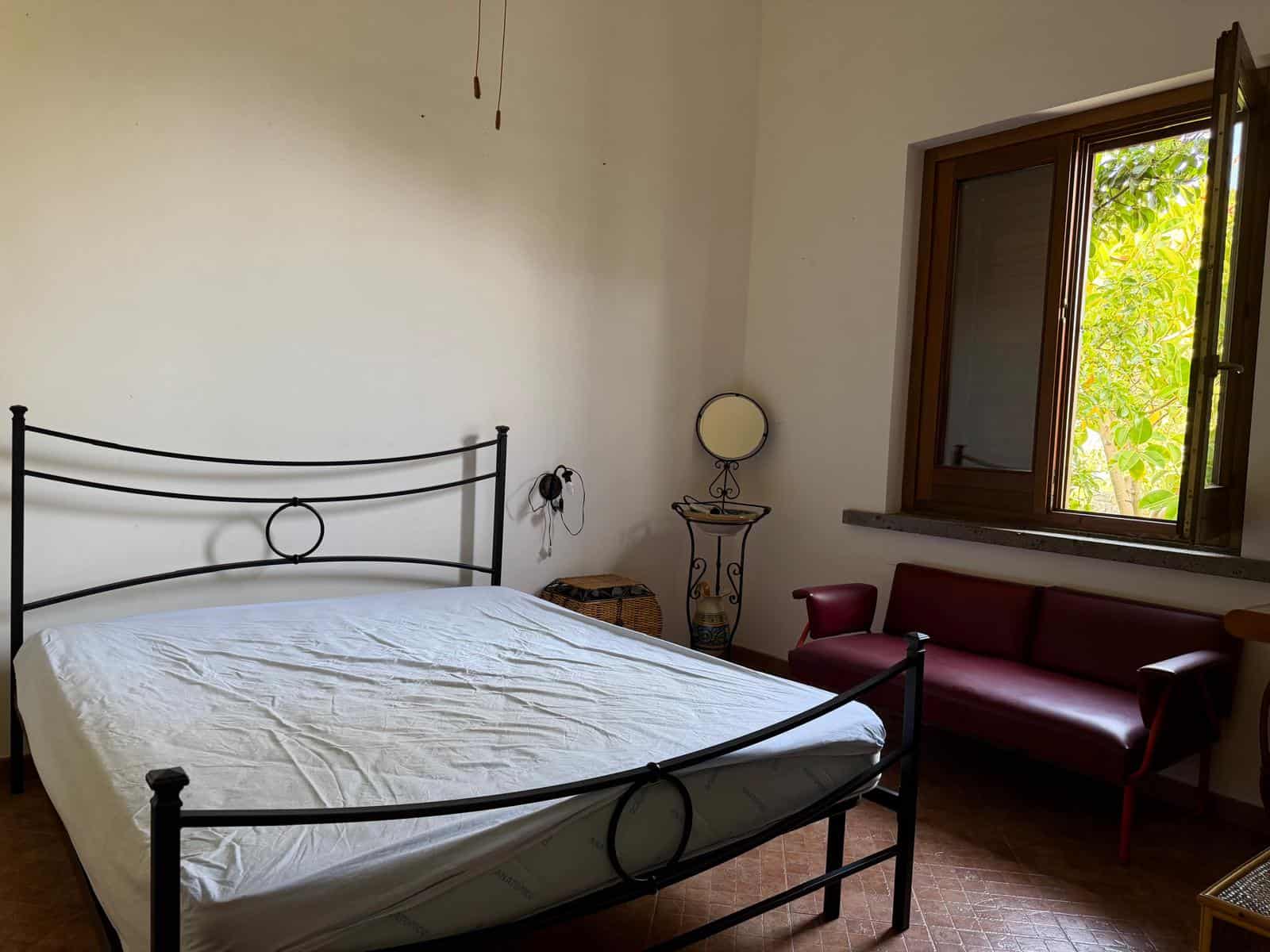 Una camera da letto luminosa con letto in ferro battuto, finestra aperta che mostra un albero verde, e arredi semplici e eleganti.
