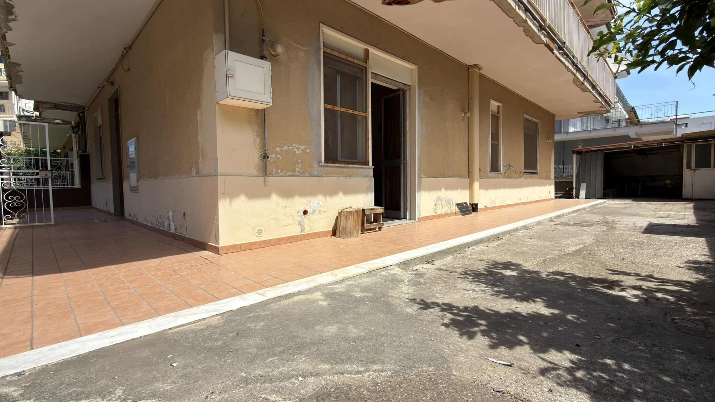 Esterno di una casa con pareti beige, finestra e porta, area di parcheggio e ombra di albero.