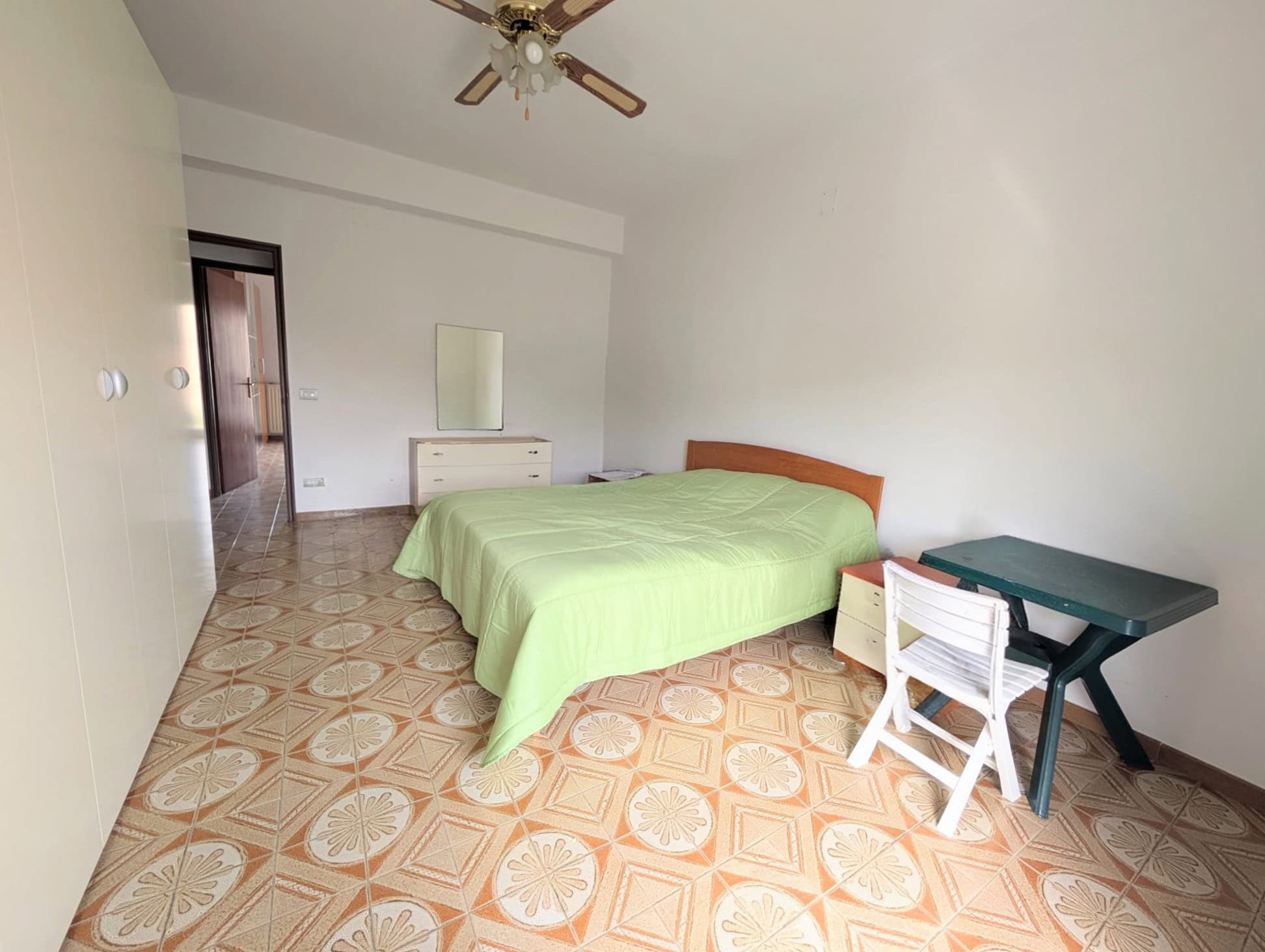 Camera che mostra una camera da letto semplice con letto, scrivania e armadio, pavimento in piastrelle decorative, pareti bianche e ventilatore a soffitto.