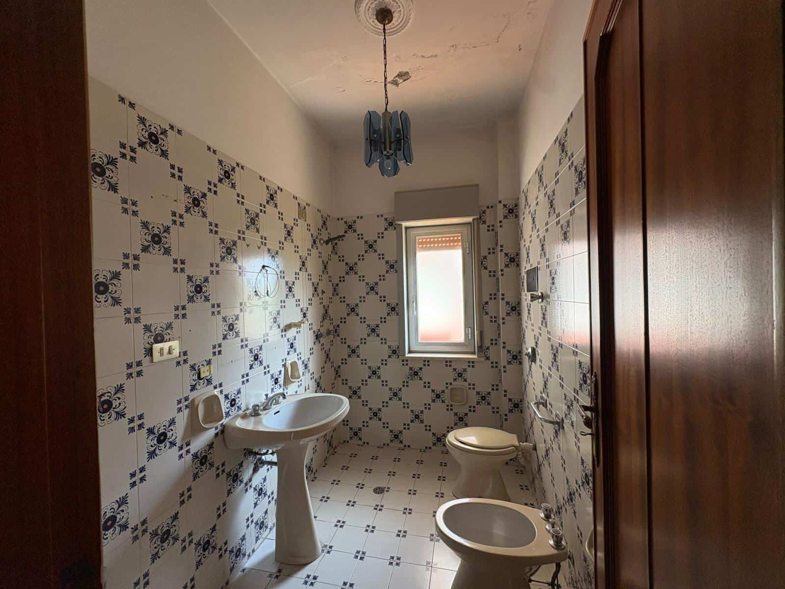 Bagno spazioso con piastrelle decorative blu e bianche, lavabo, WC e bidet, finestra naturale, illuminazione centrale, in un appartamento in vendita a Merì.
