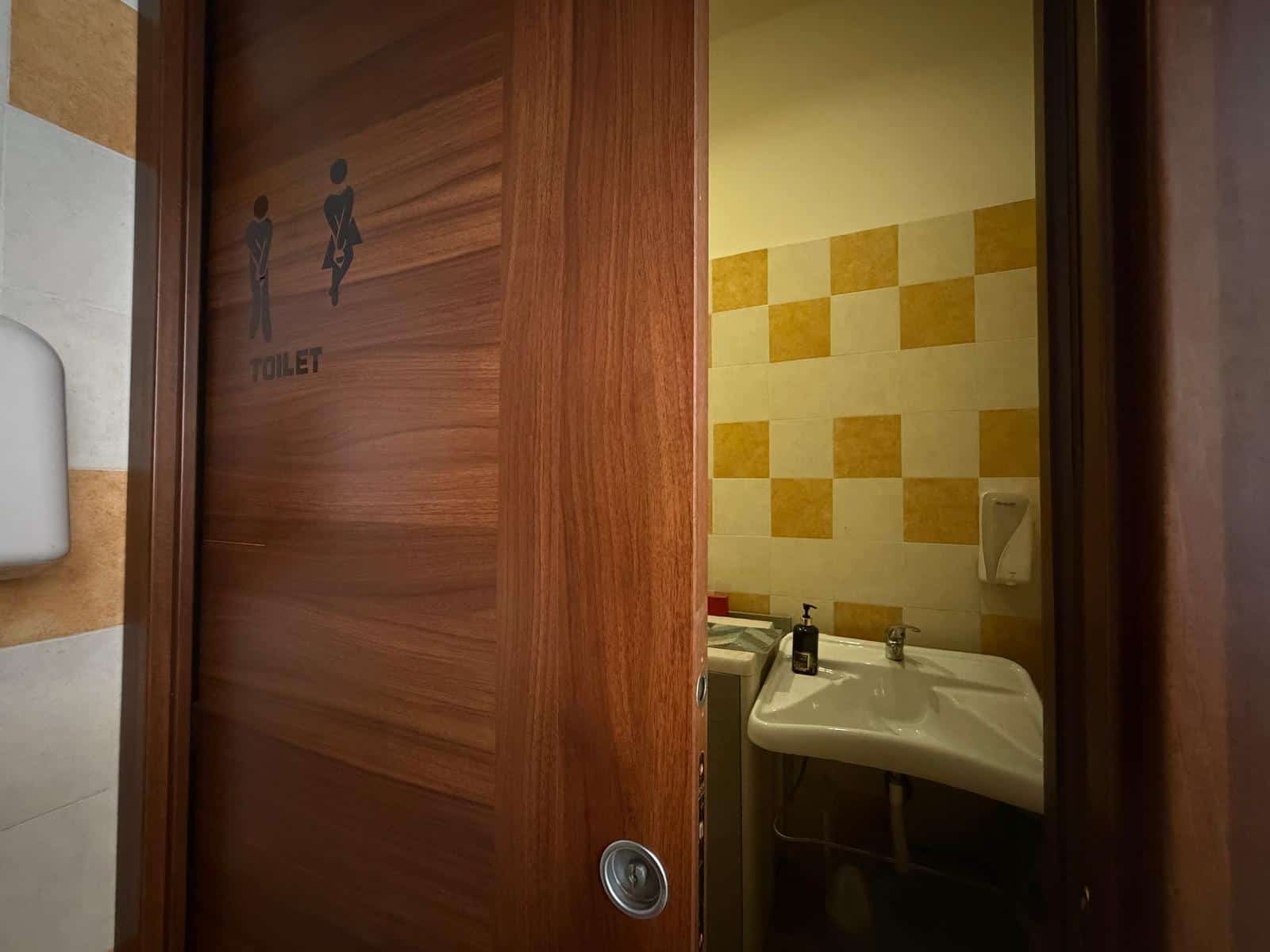 Bagno pubblico con lavabo e pareti decorate con piastrelle gialle e beige, accessibile tramite porta in legno.