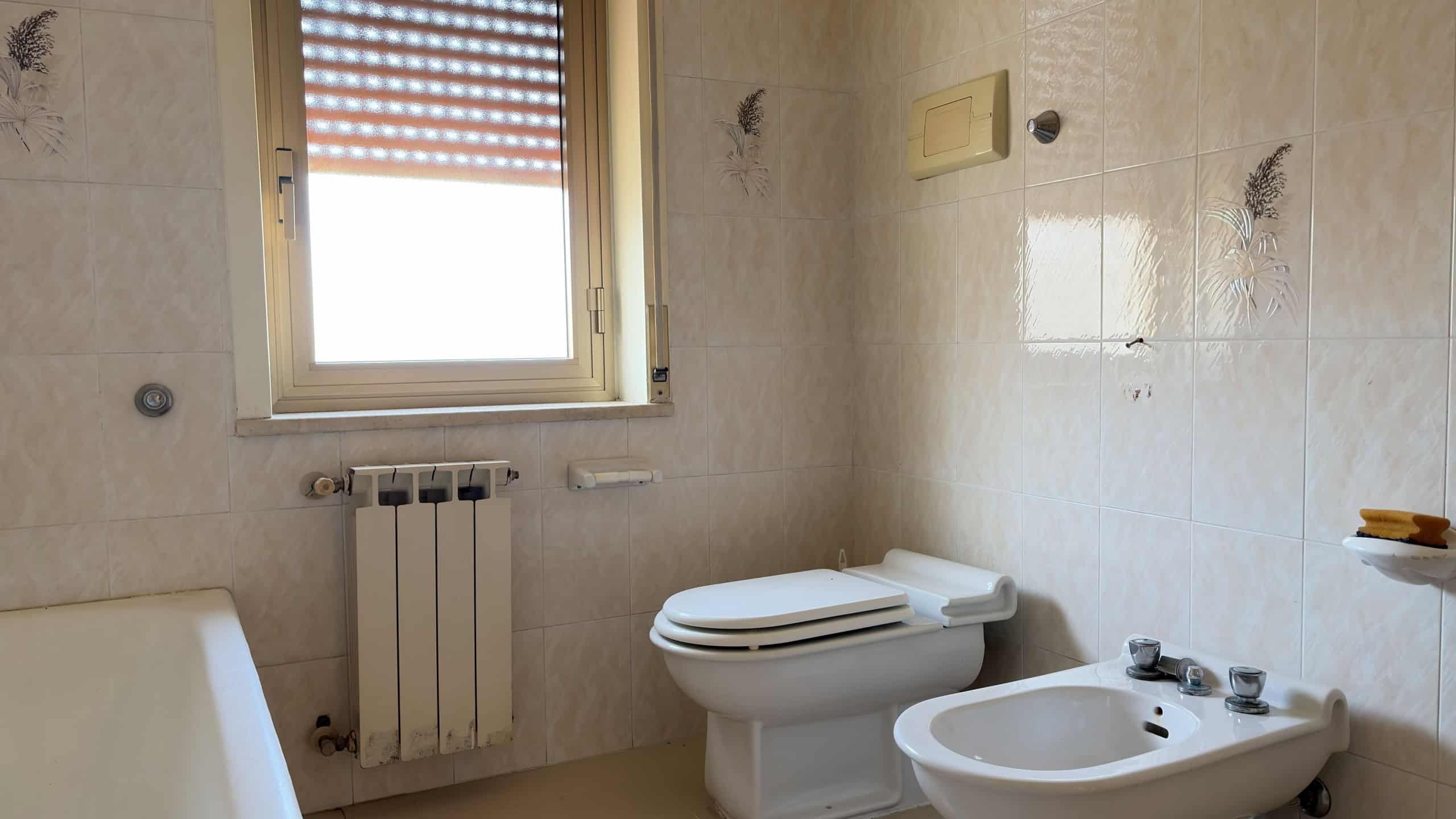 Bagno con pareti rivestite in piastrelle beige, finestra con tapparella, radiatore e servizi igienici moderni.