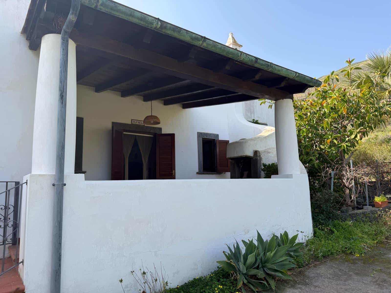 Casa tradizionale in stile mediterraneo con veranda e piante verdi, ideale per case vacanza o residenze estive, immersa nel verde e con un'architettura accogliente.