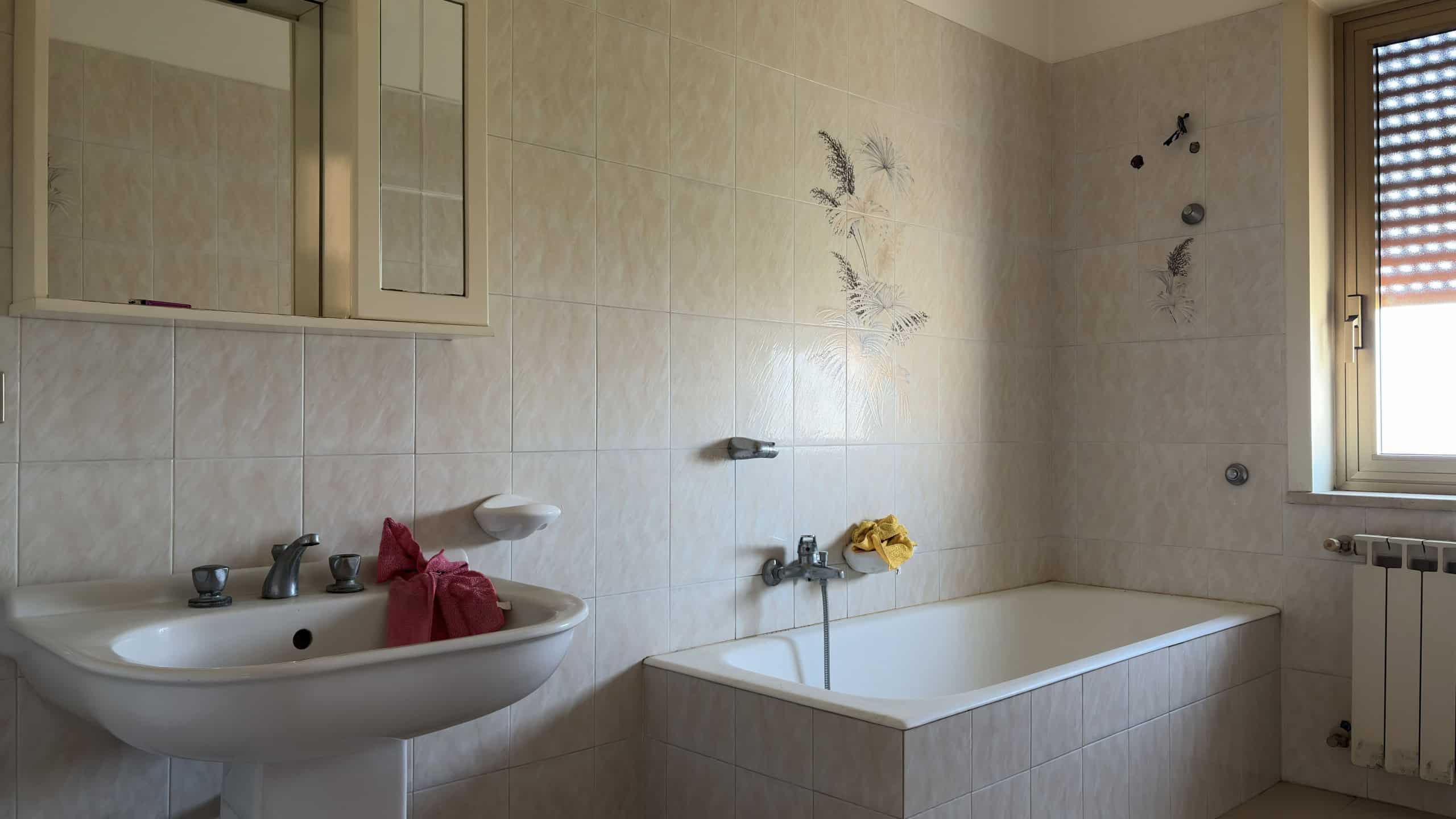 Bagno con vasca e lavabo in un ambiente luminoso e pulito, finestra con persiane e pareti beige.
