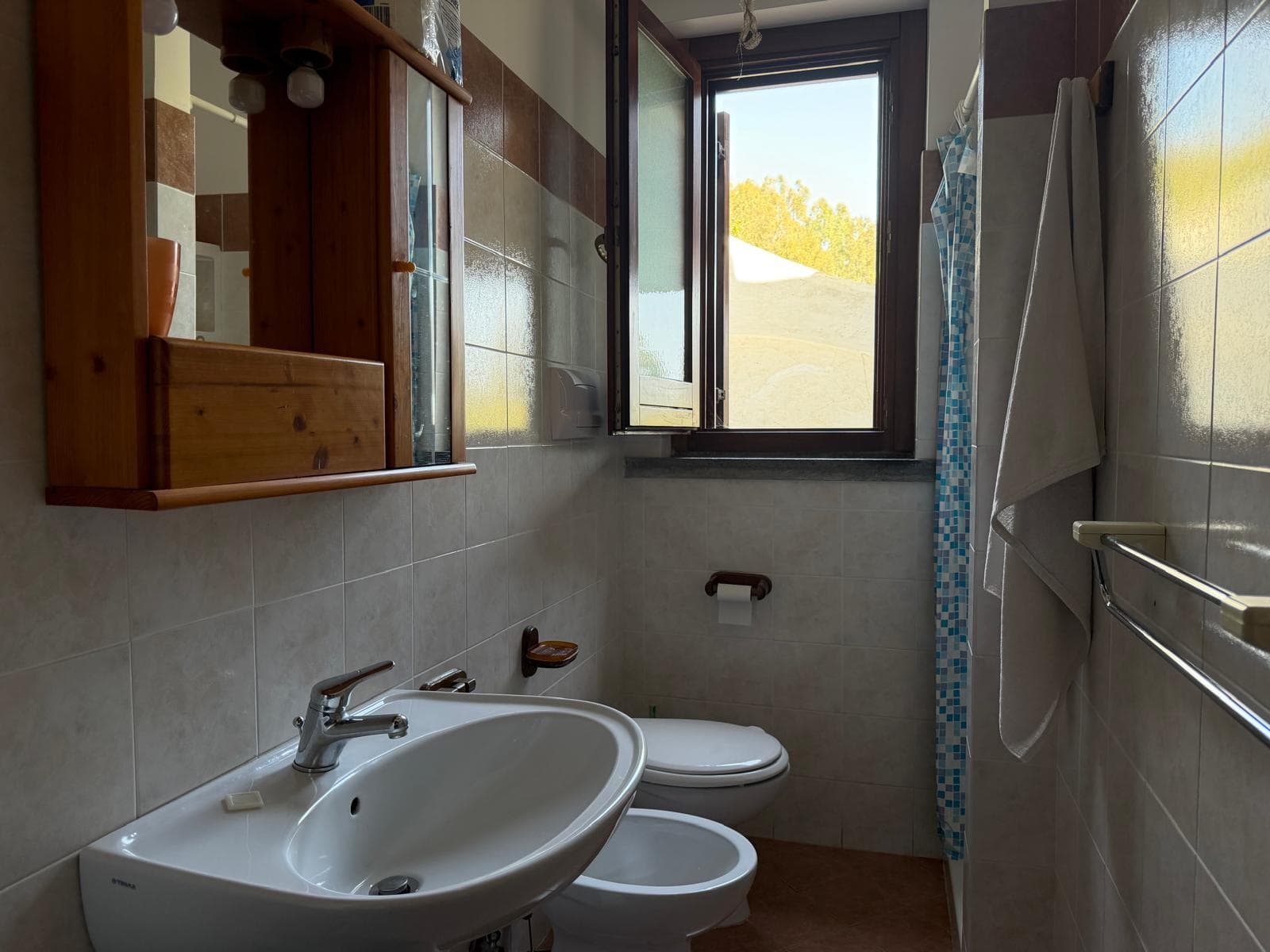 Lavabo bianco con rubinetto in metallo, specchio e armadietto in legno, finestra aperta che mostra un paesaggio naturale, in bagno luminoso e funzionale.
