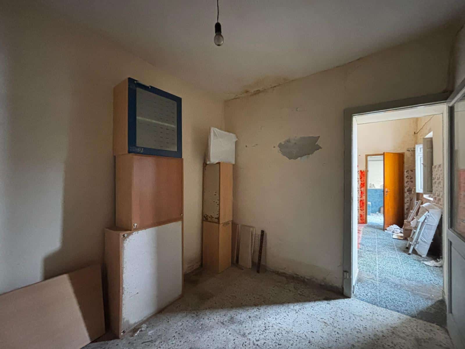 Il testo dell'immagine mostra una stanza vuota in fase di ristrutturazione, con pareti danneggiate e materiali da costruzione accatastati.
