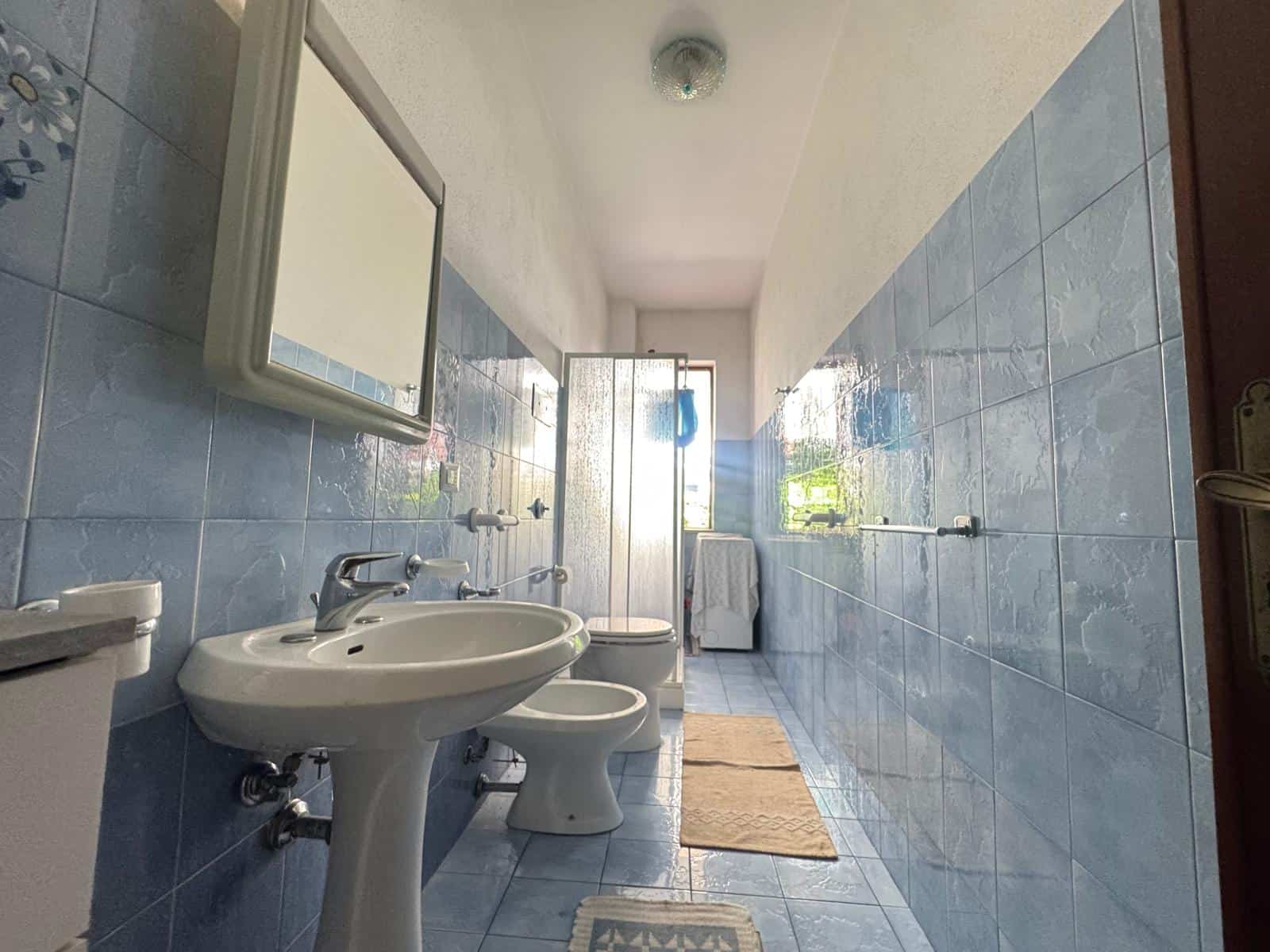 Lavabo e servizi igienici in bagno con piastrelle azzurre, finestra luminosa e asciugatrice, ambiente spazioso e luminoso, ideale per proprietà in affitto o vendita a NextCasa.