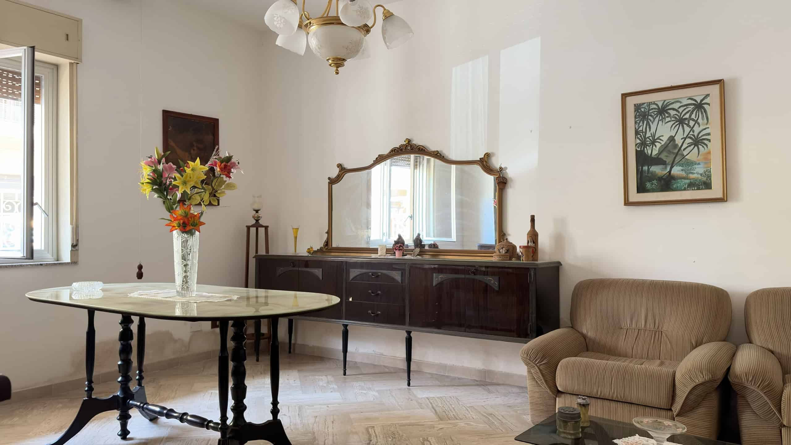 Interno di un soggiorno con mobili d'epoca, grande specchio e decorazioni floreali, ambiente luminoso e accogliente.