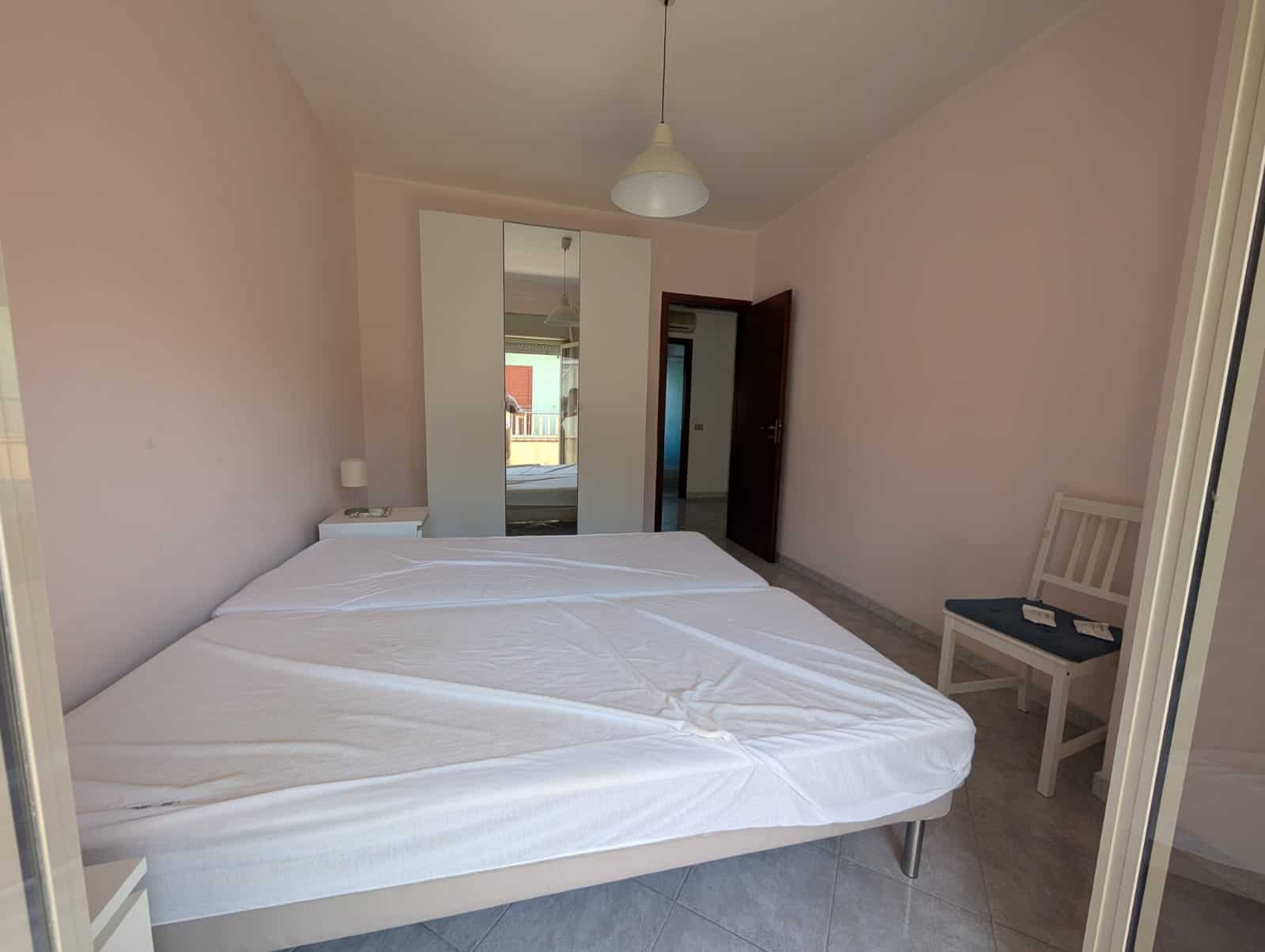 Letto matrimoniale in camera da letto con pareti rosa chiaro, armadio a specchio e sedia bianca, ambiente luminoso e semplice, ideale per affitti turistici o residenziali.
